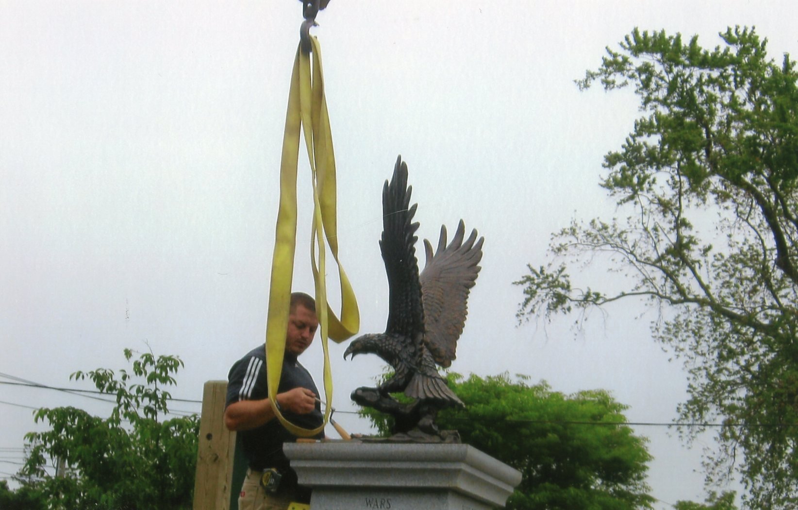 A man working on a sculpture of a bird with outstretched wings, hanging from a yellow strap, outdoors with trees in the background.