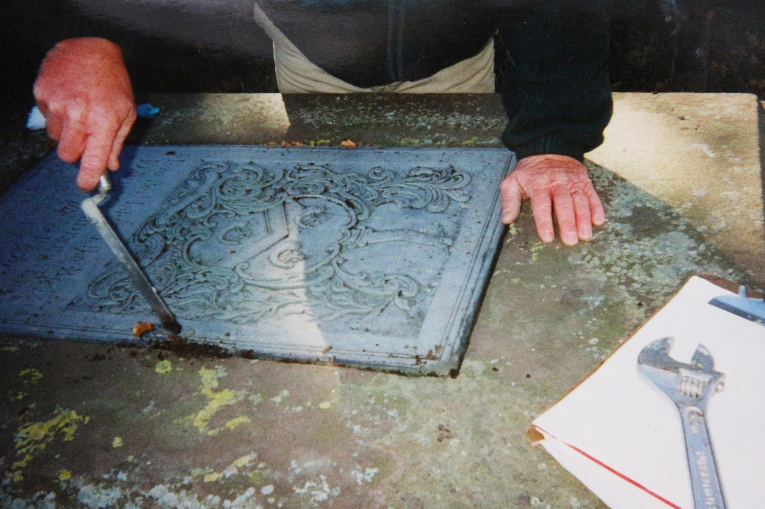 A person working on an ornate metal plaque outdoors, with hands visible, one holding a tool and the other resting on the plaque. The person is wearing a dark jacket and a light-colored apron, and the setting appears to be a workshop or outdoor worksp