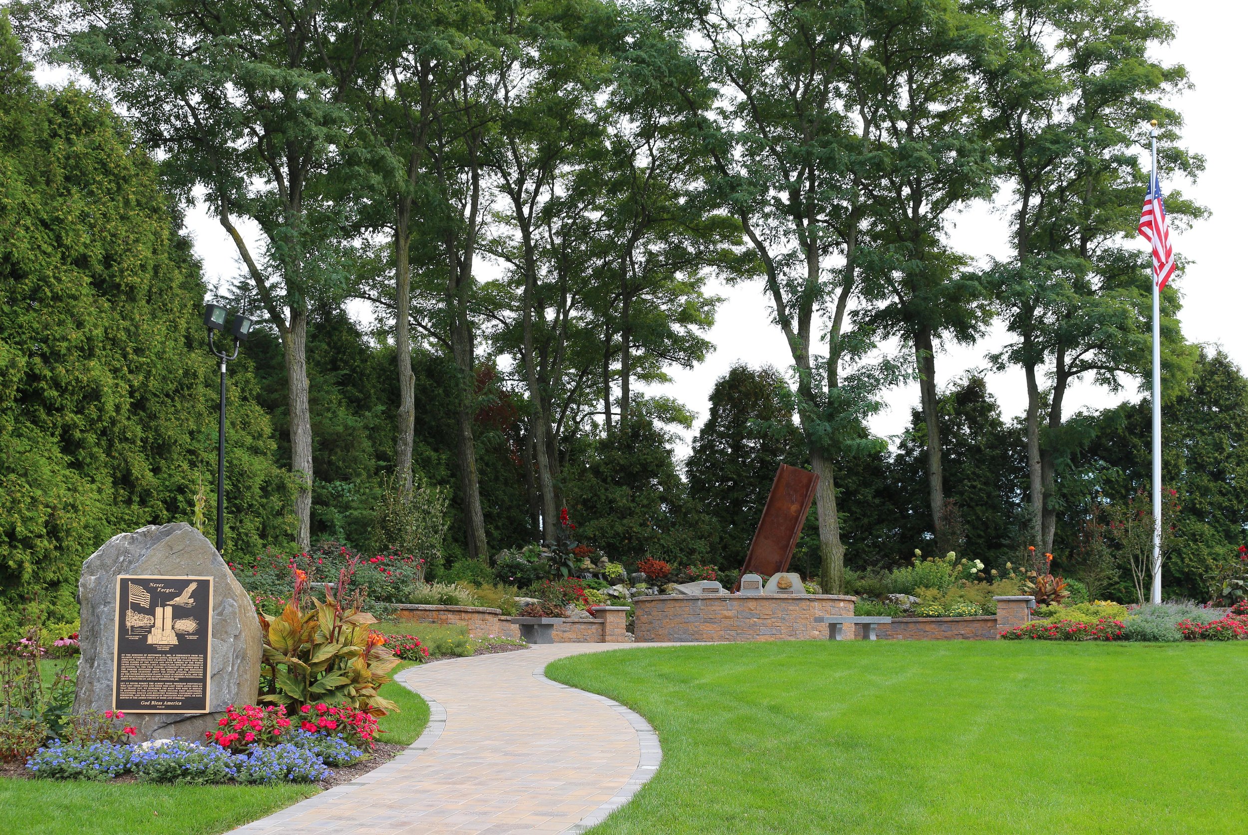 A landscaped garden with a brick pathway curving through green grass, surrounded by colorful flowers, trees, a memorial plaque, and an American flag on a flagpole.