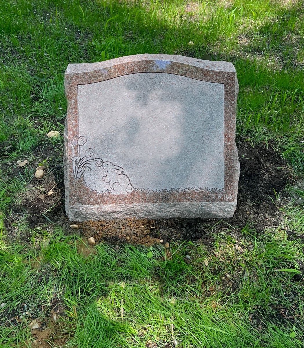A gray tombstone with decorative flower engravings at the bottom left corner, situated in a grassy area with patches of dirt around it.