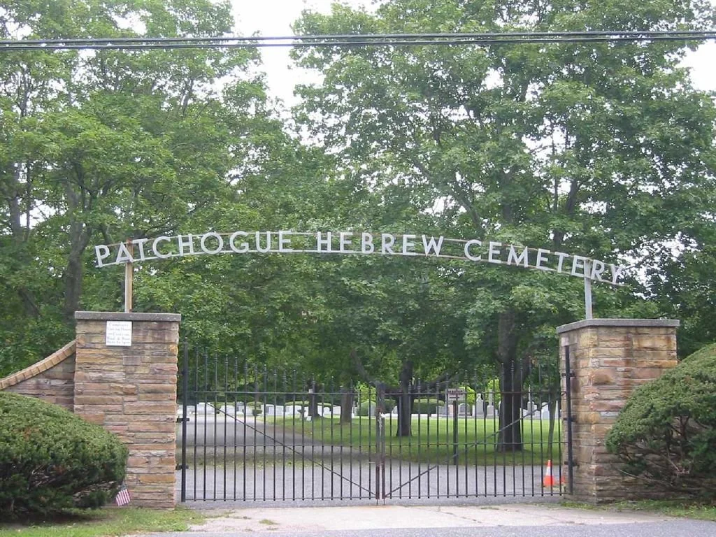Entrance gate to Patchogue Hebrew Cemetery, with a black metal gate, stone pillars, and a sign arching overhead, surrounded by green trees and bushes.
