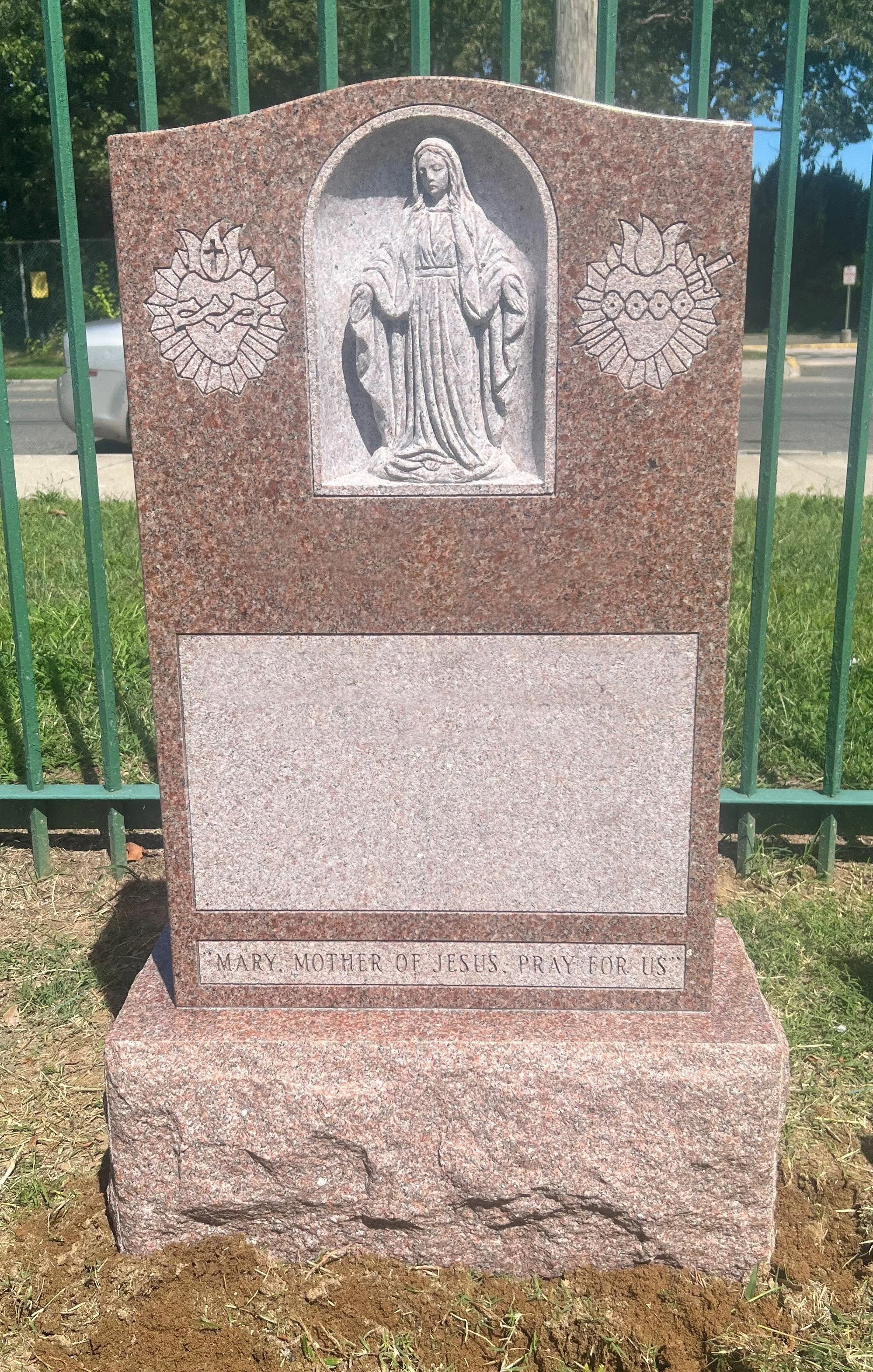 A gravestone with a carved image of Mary, mother of Jesus, praying. The gravestone has a pinkish granite appearance and the inscription: "Mary, Mother of Jesus, Pray for Us."