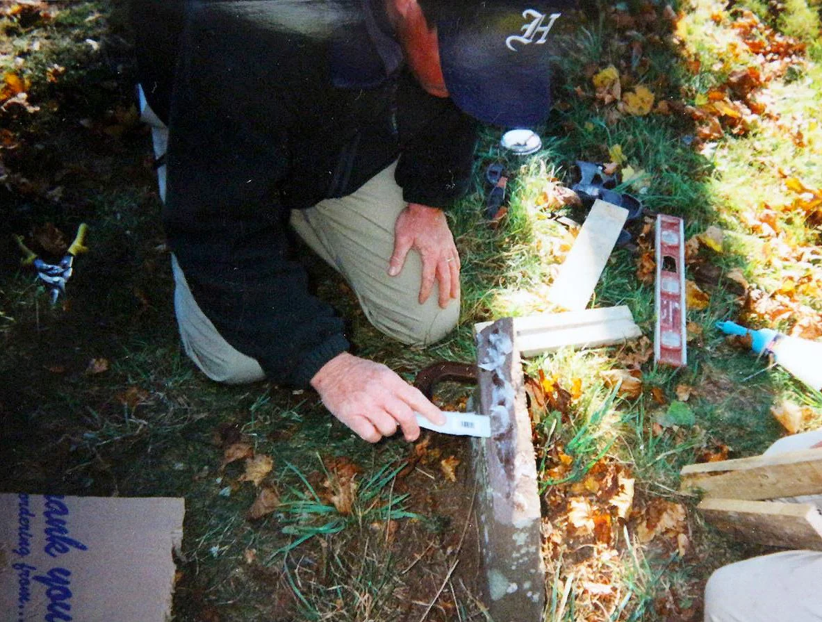 Person kneeling on the ground outdoors working with tools, surrounded by leaves and grass.