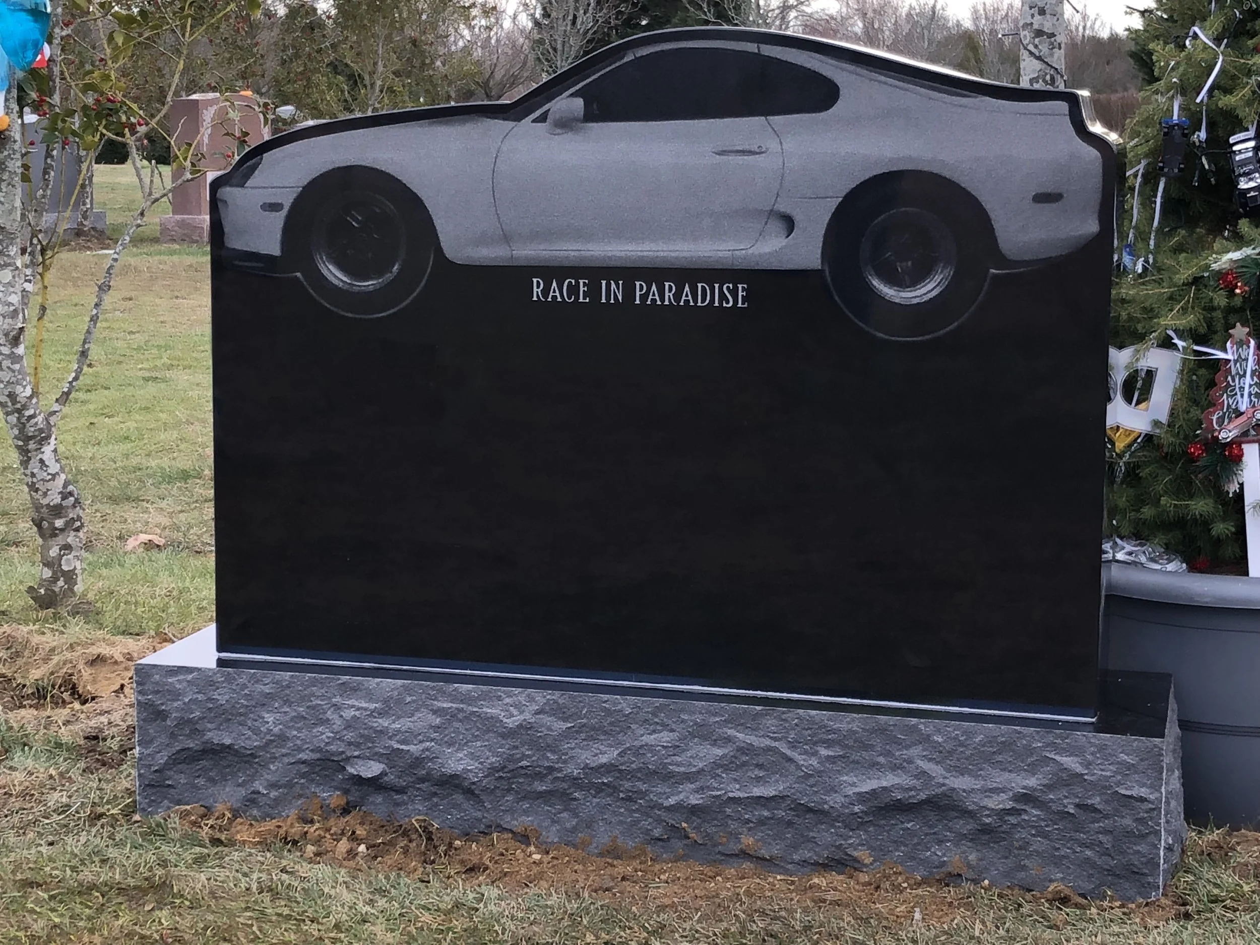 Granite monument with an image of a racing car and the words "Race in Paradise" engraved on it, situated outdoors with trees and holiday decorations nearby.