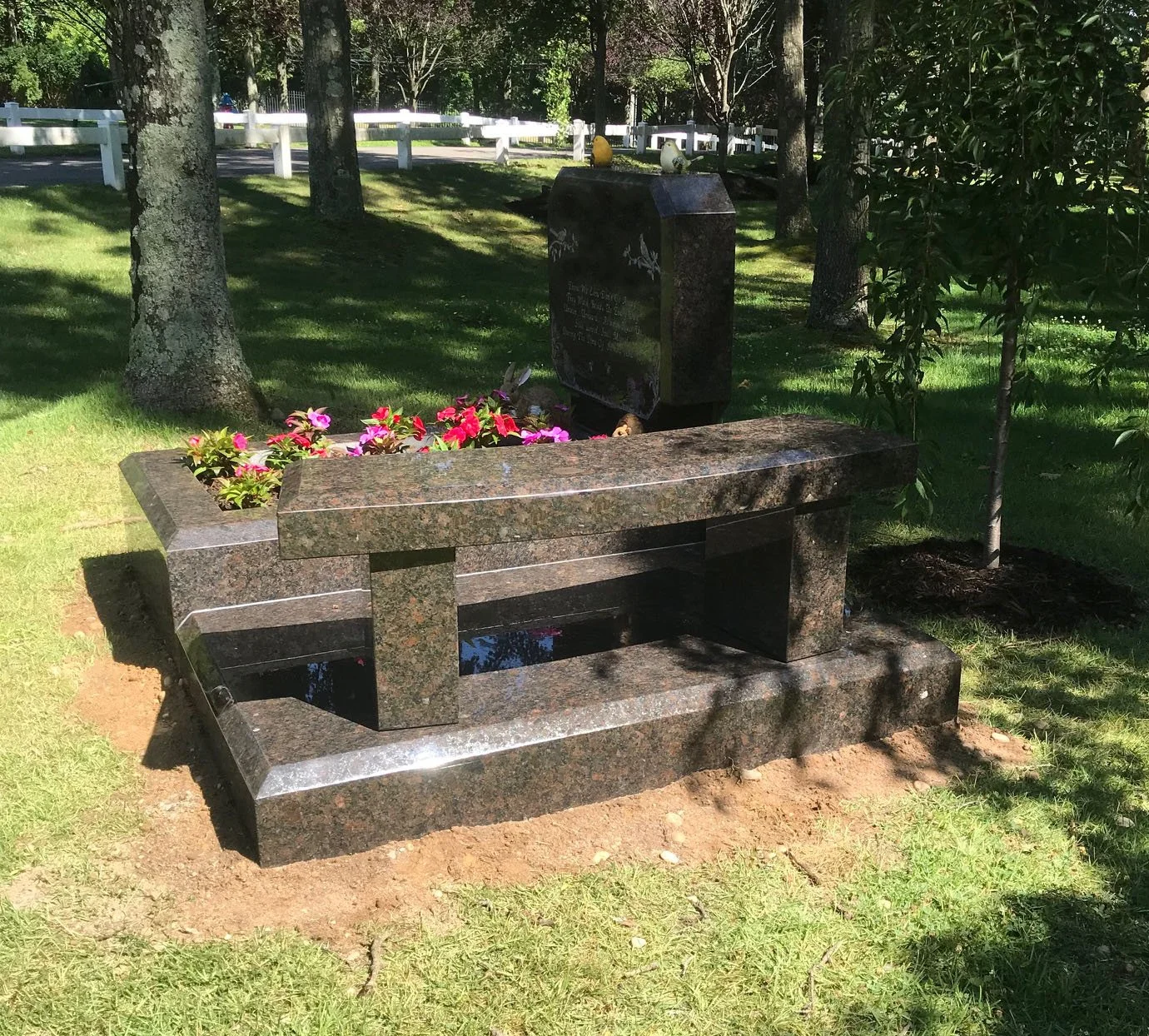 A granite memorial bench with a flower bed on one end, located in a grassy park with trees and a white fence in the background.