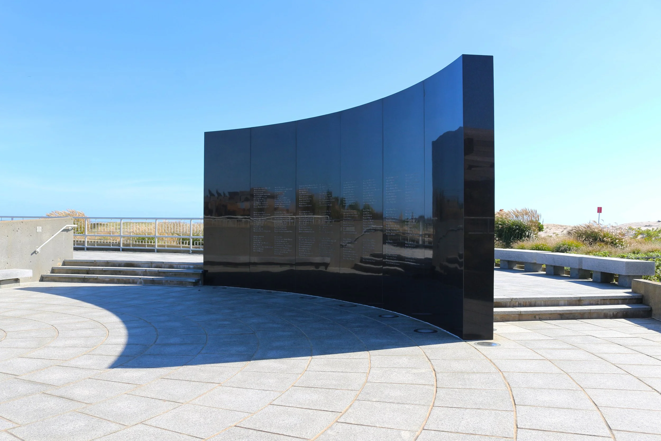 A black memorial wall with engraved names, positioned outdoors on a sunny day. In the background, there are grassy dunes, a clear blue sky, and a white railing.