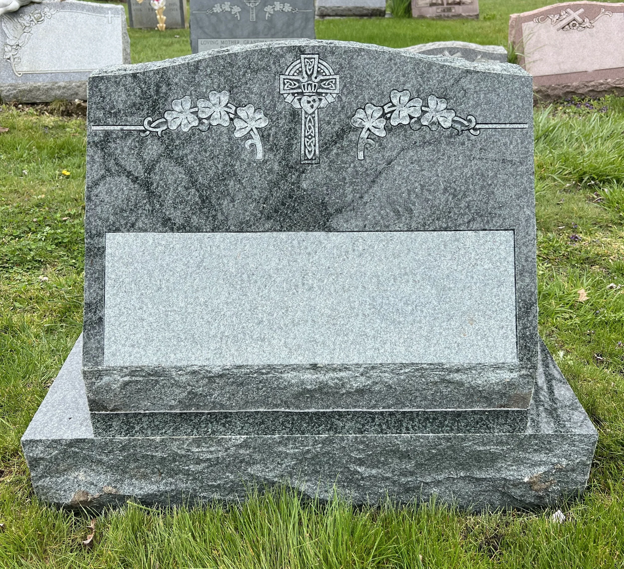A gray granite headstone with engraved floral and Celtic cross designs, set in a grassy cemetery, with other headstones in the background.