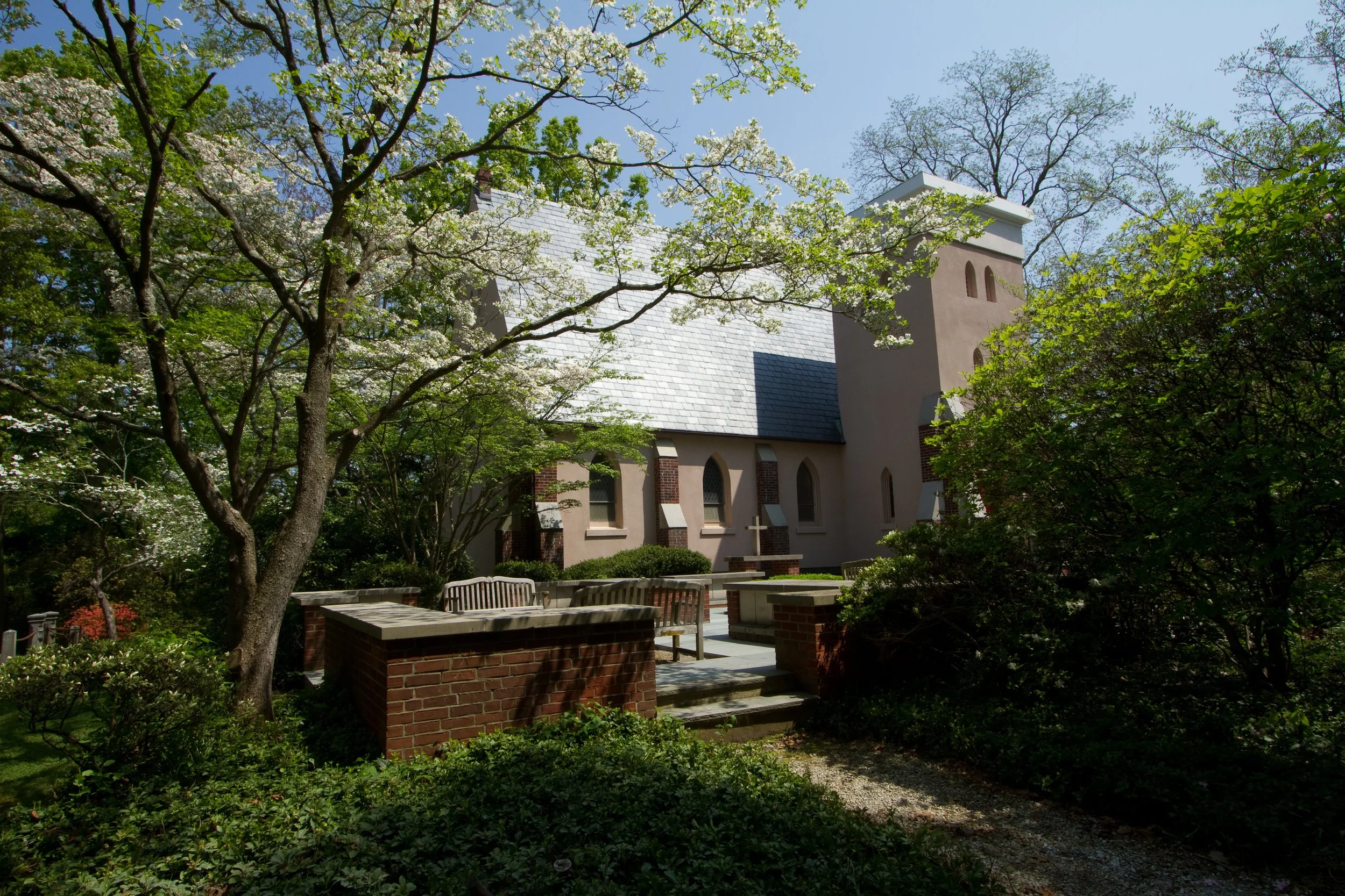 A church surrounded by trees and greenery, with a light-colored exterior and a sloped roof, partially obscured by blossoming tree branches, sunlight filtering through the leaves.