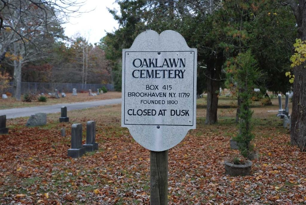 Sign in Oaklawn Cemetery indicating it is closed at dusk, with trees and gravestones in the background.