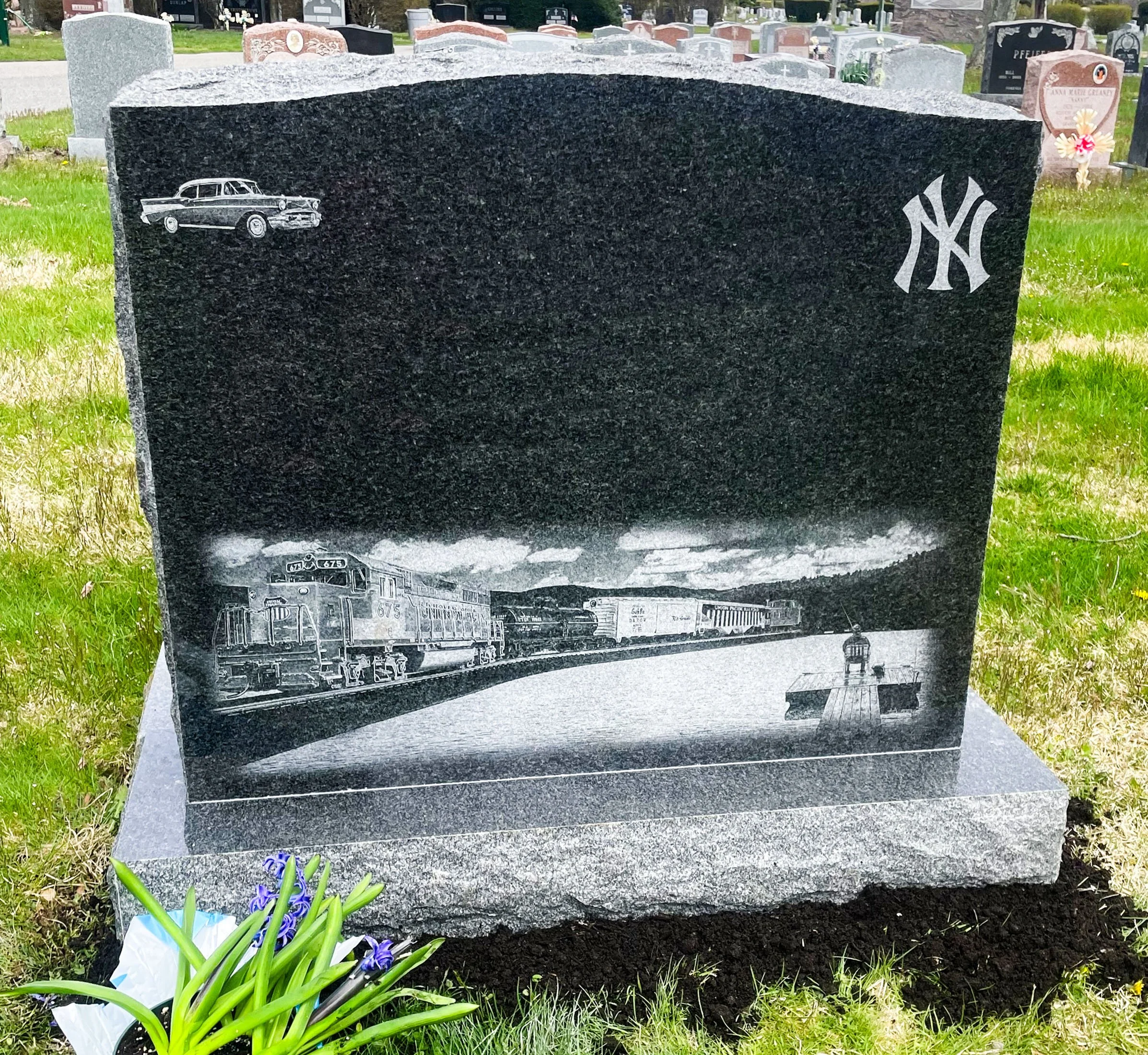 A black polished granite headstone in a cemetery with engraved images of a vintage car and a train, and the New York Yankees logo. There are yellow flowers and purple irises at the base of the headstone, with other graves and monuments visible in the background.