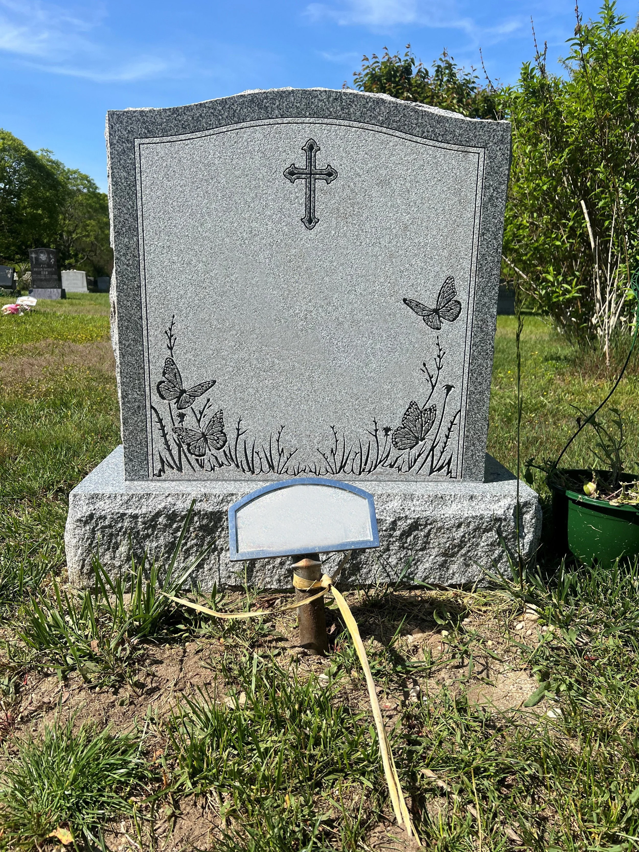 Gravestone with a cross at the top, decorated with butterflies and grass designs, in a cemetery under a blue sky.