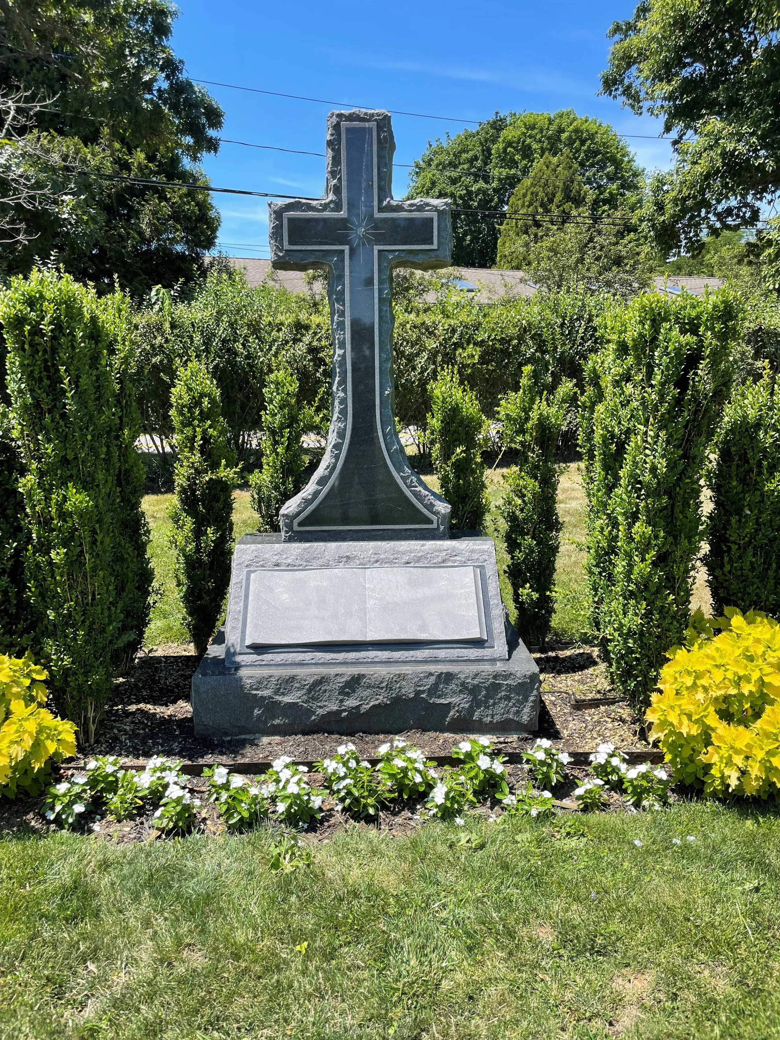 A stone cross grave monument surrounded by green shrubs and yellow and white flowers under a bright blue sky.