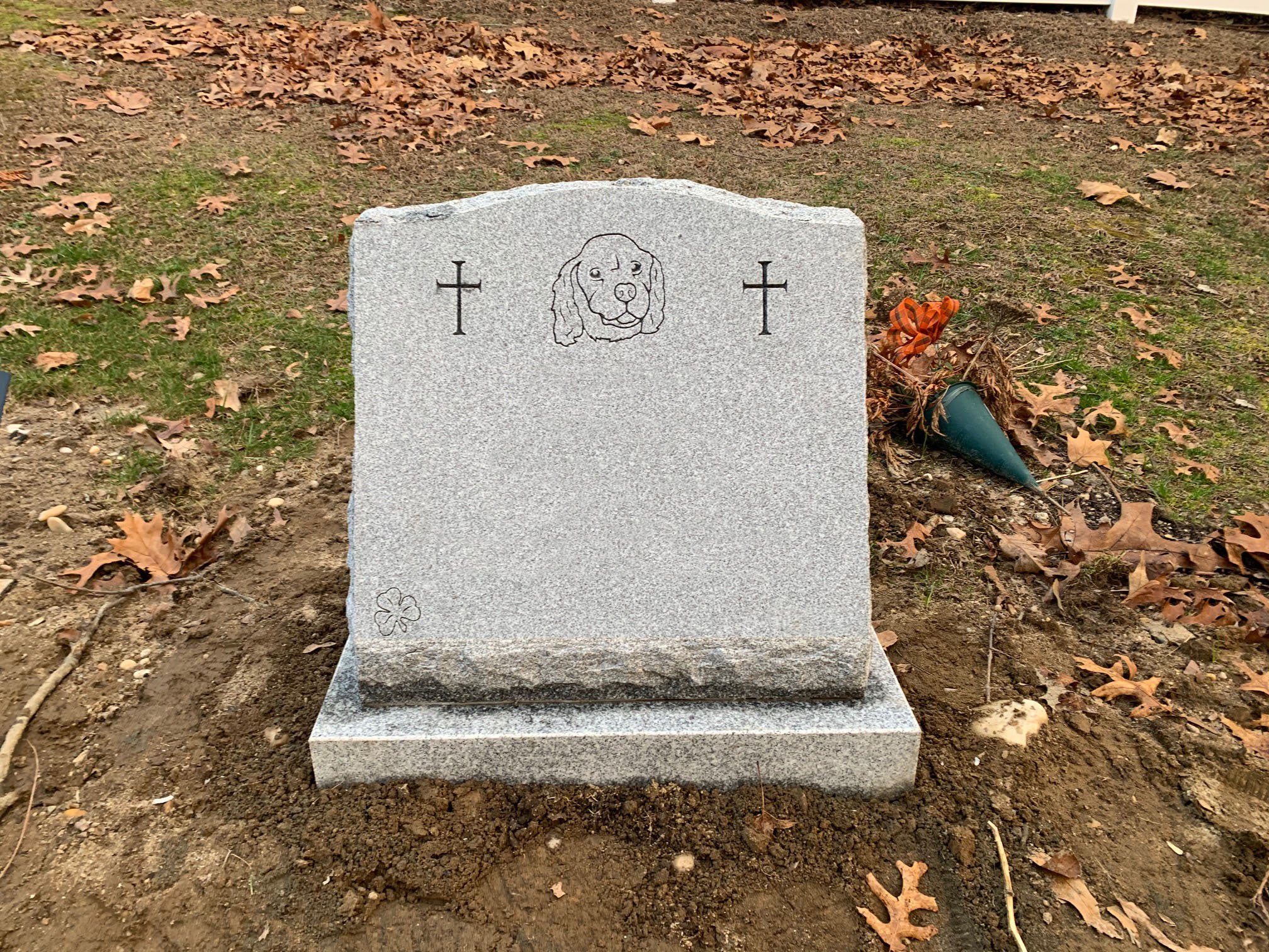 Gray granite headstone with a carved image of a dog's face, flanked by two crosses, on a bed of dirt surrounded by fallen leaves.