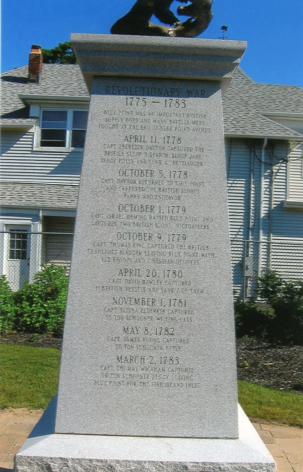 Historical monument with engraved text about Revolutionary War events, located in front of a house with a gray roof and white siding, surrounded by grass and a chain-link fence.