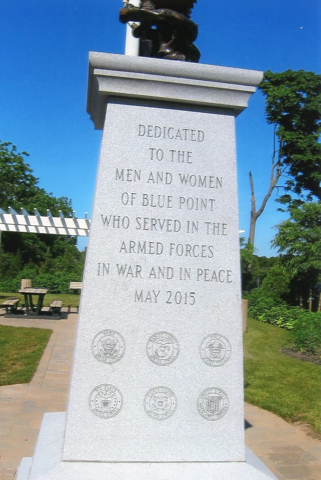 A stone monument dedicated to the men and women of Blue Point who served in the armed forces, located outdoors with trees, benches, and a picnic table in the background, under a clear blue sky.
