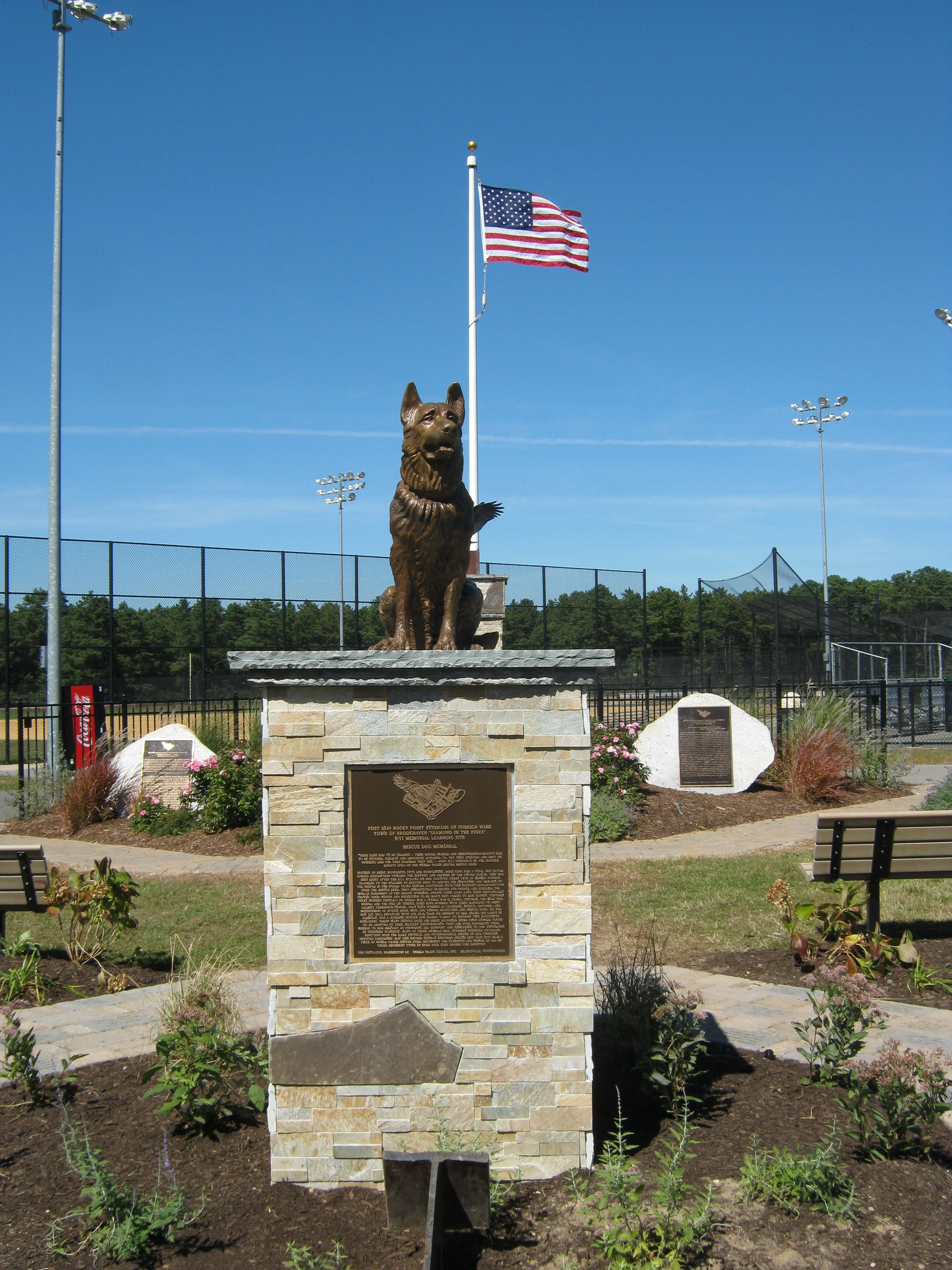A bronze statue of a dog sitting on a stone pedestal, with a plaque, in a landscaped area. Behind it is a flagpole with an American flag, and a sports field with floodlights. The sky is clear and blue.