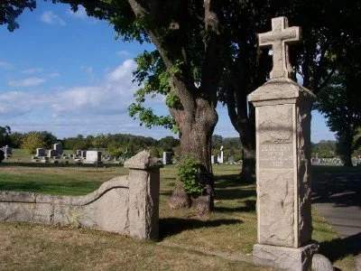 A stone cemetery entrance with a cross on top, surrounded by trees and gravestones in the background.