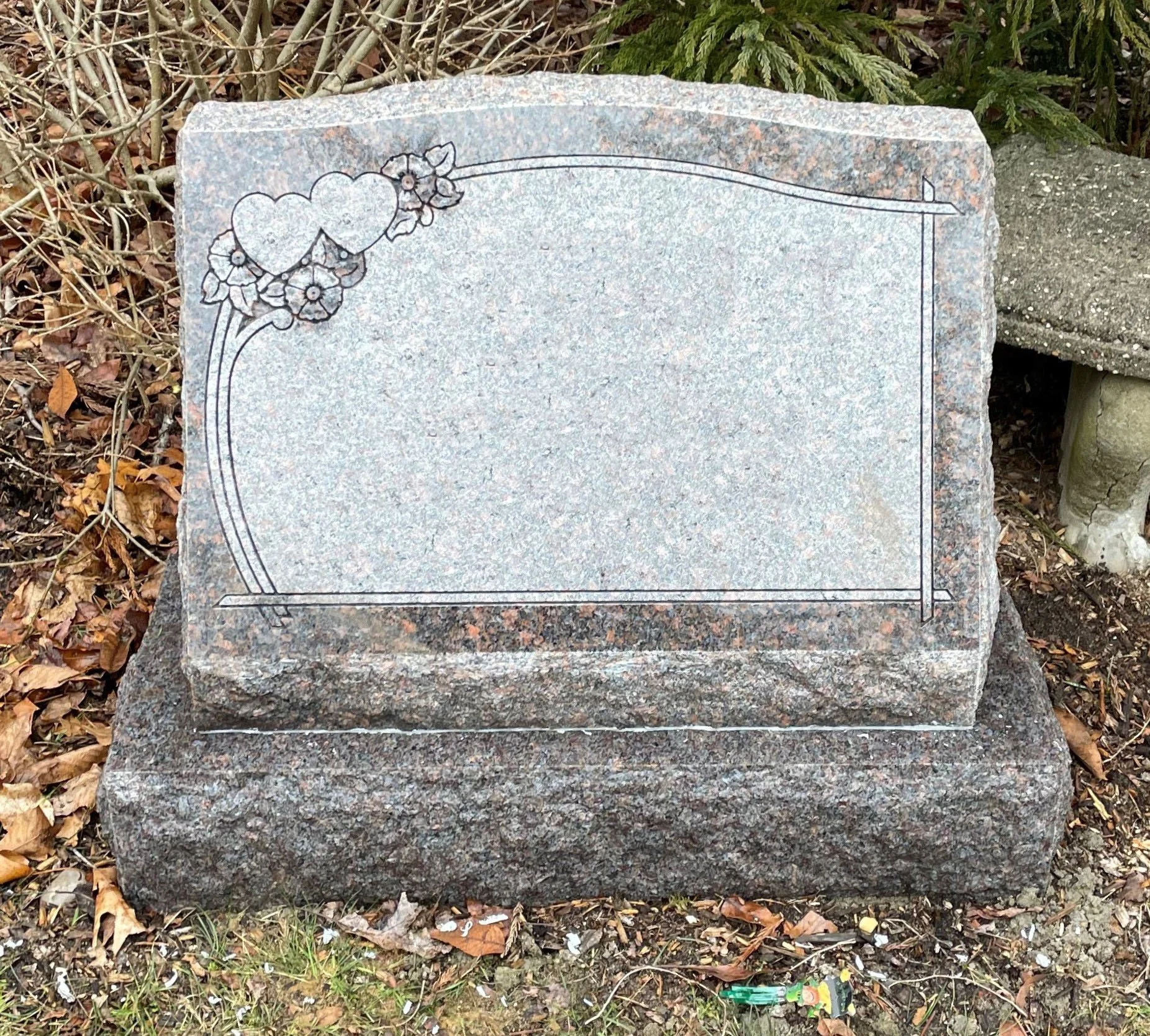 A gray granite tombstone with hand-etched floral border design, set outdoors among fallen leaves and shrubbery.
