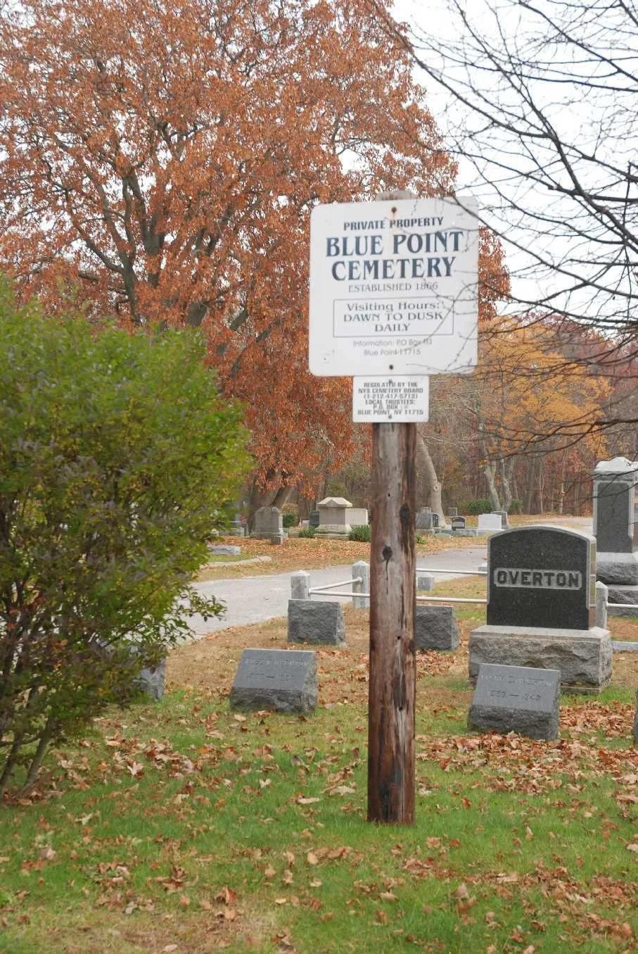 A cemetery with a wooden sign that reads 'Blue Point Cemetery', established in 1886, with visiting hours from dawn to dusk daily. Tombstones and trees with autumn leaves are visible in the background.