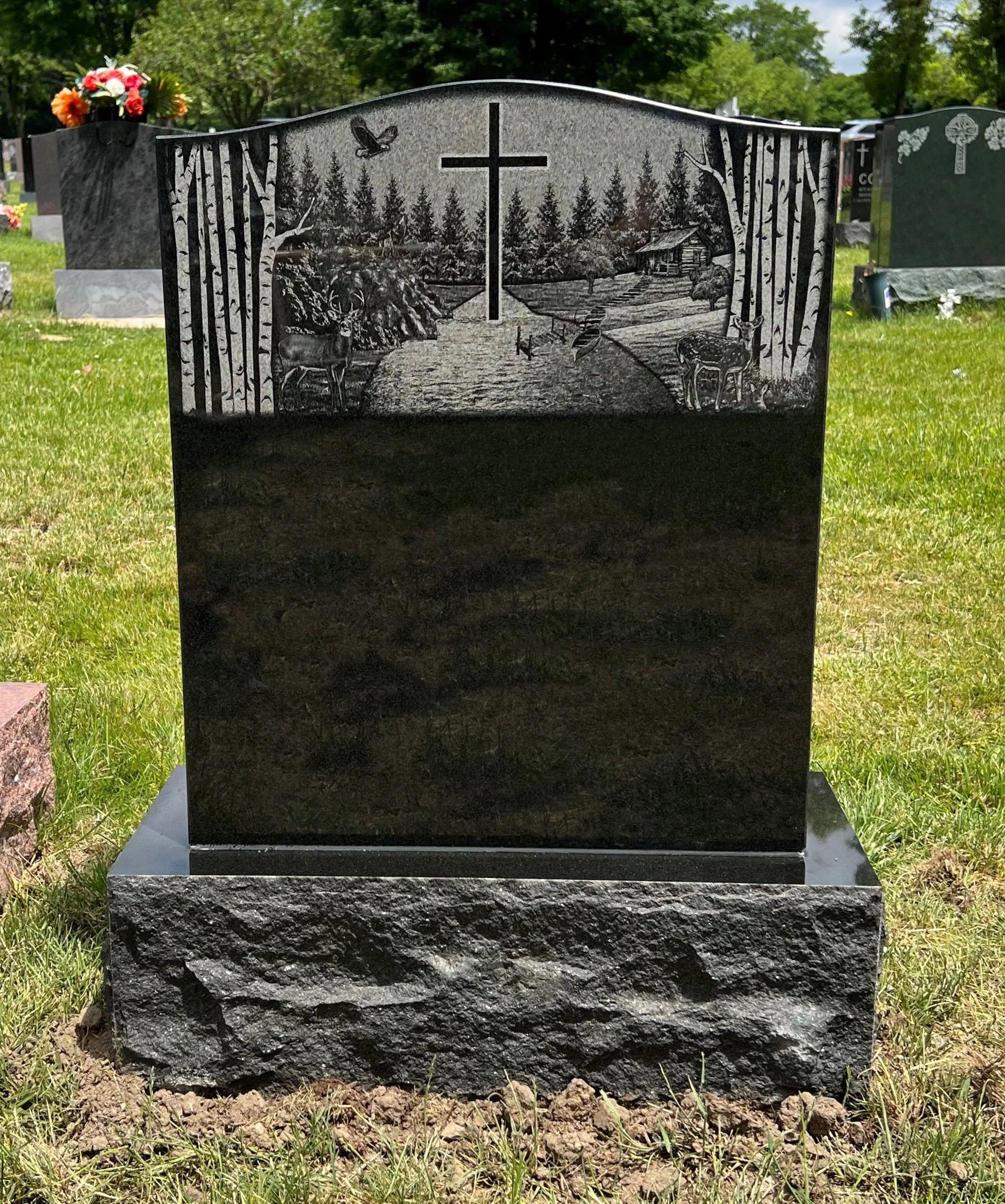 A black granite headstone with a detailed engraving of a rural landscape, including a log cabin, trees, deer, and birds, set in a grassy cemetery.