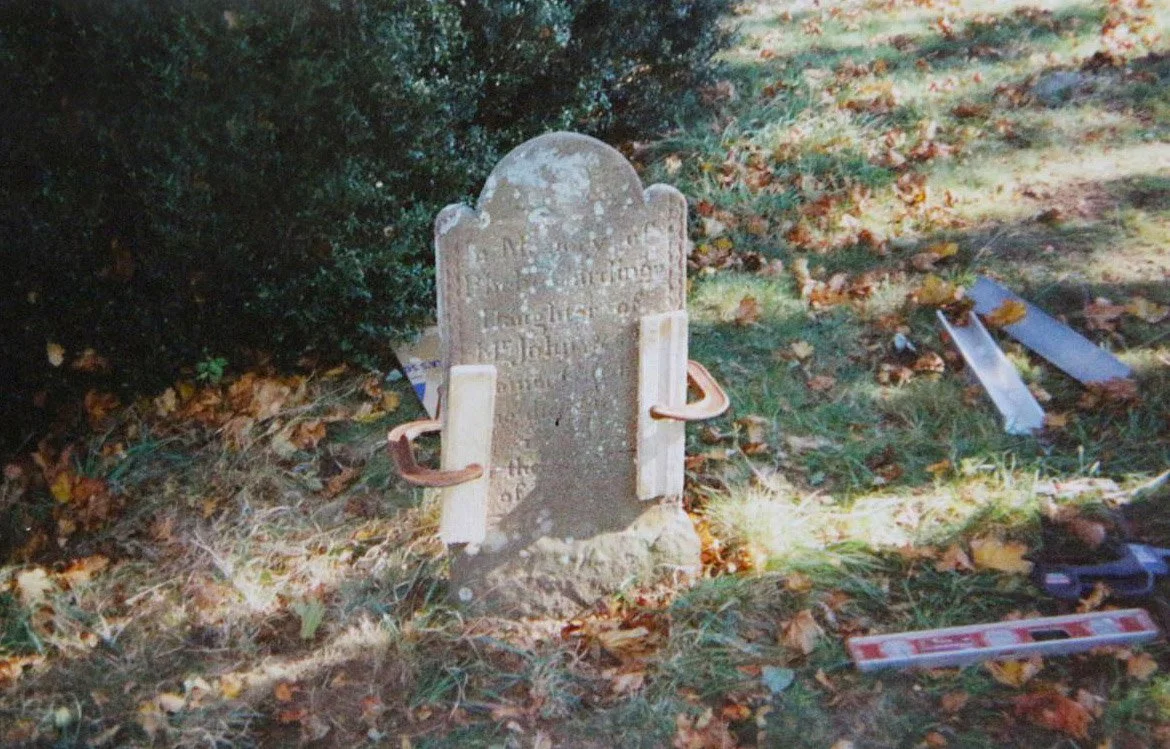 Old, weathered tombstone in a grassy area surrounded by fallen leaves, with tools and objects scattered nearby.