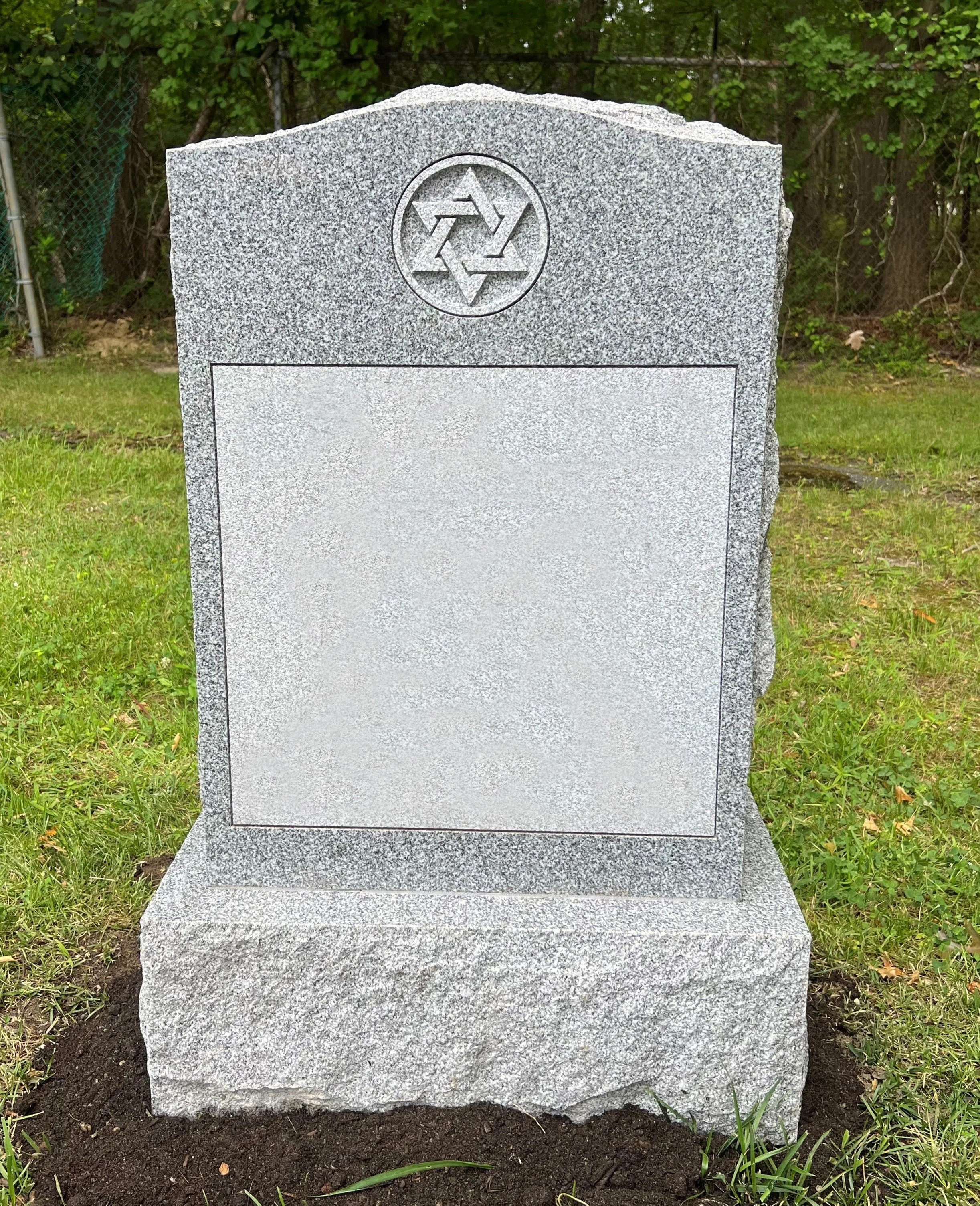 Gray granite headstone with a Star of David symbol engraved at the top, standing upright on a bed of soil, surrounded by green grass and trees in the background.