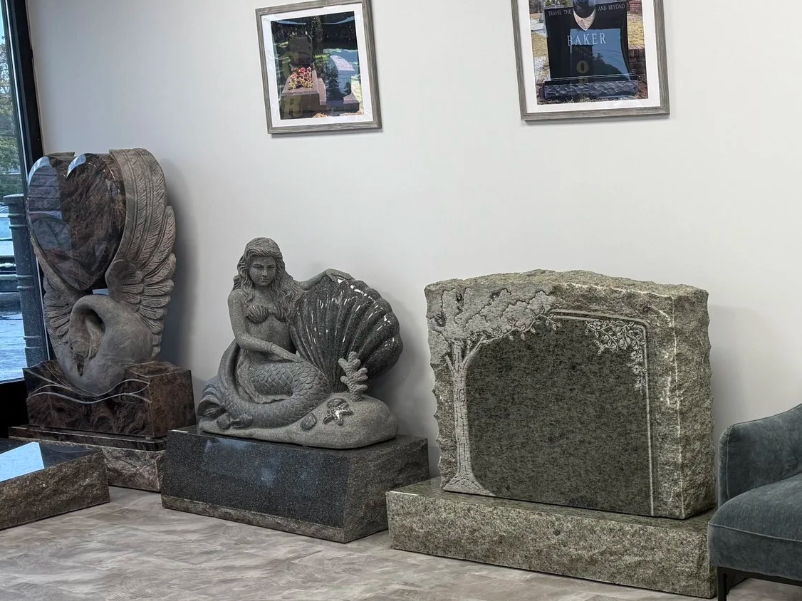 Group of three large sculptures and a framed picture on the wall. The sculptures include a heart-shaped sculpture with swan and wave details, a mermaid sculpture holding a large shell, and a rectangular carved stone with floral design. Part of a gray upholstered chair is visible on the right.