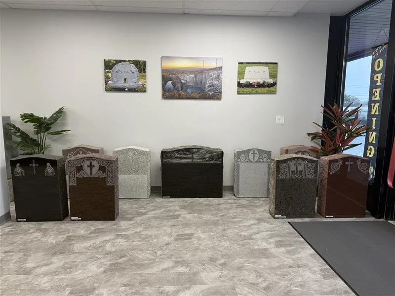 A display of eight polished granite headstones arranged in a row inside a showroom, with three landscape wall art pieces above them and two potted plants on either side.
