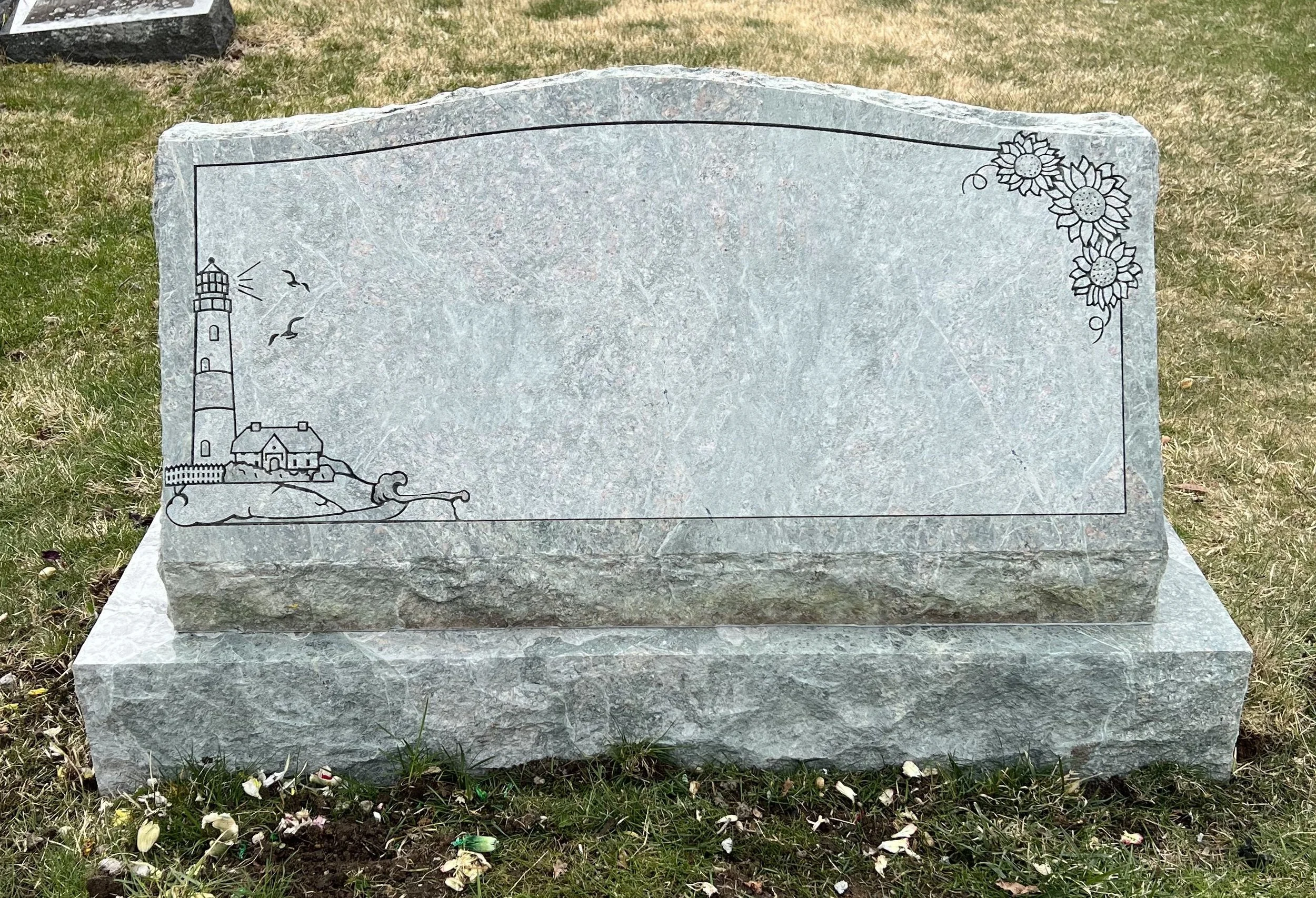 A gray stone grave marker with an engraved illustration of a lighthouse, some birds, and flowers, situated on a grassy area.