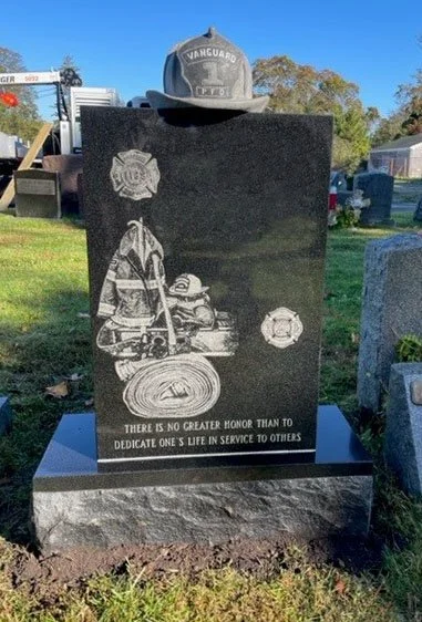 A black granite tombstone with etched images of a firefighter and firefighter equipment, a firefighter hat on top, and an inscription that reads "There is no greater honor than to dedicate one's life in service to others."