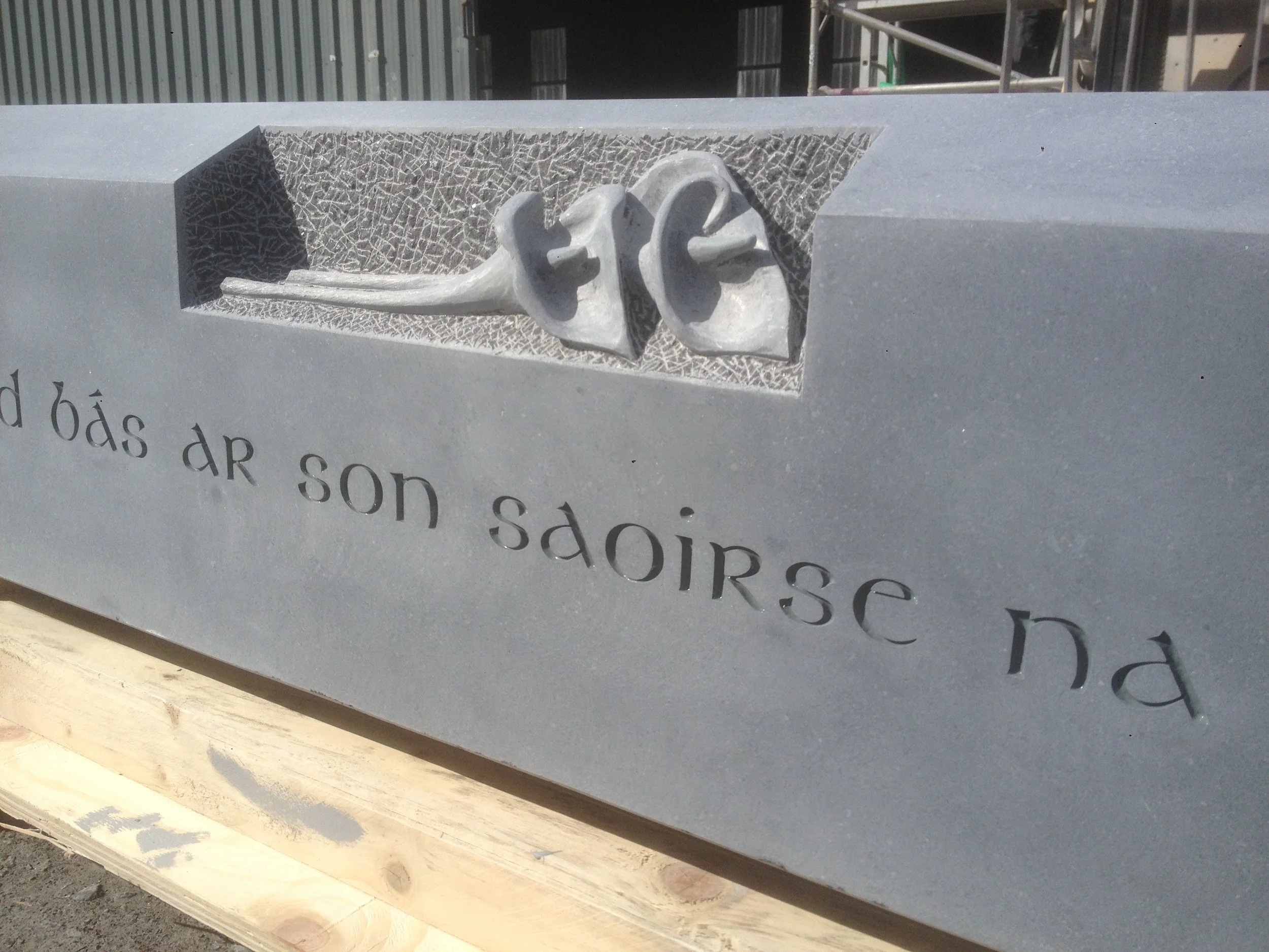 Close-up view of a memorial plaque with a sculpted rose symbol in metal, mounted on a stone or concrete surface, with partially visible engraved text.