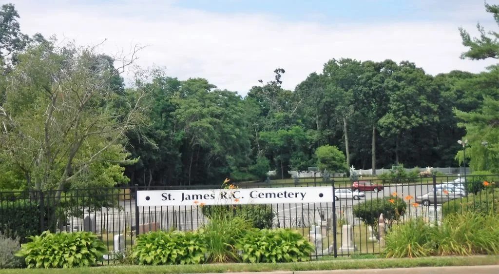 Entrance to St. James R.C. Cemetery with a sign, stone grave markers, and a parking lot surrounded by trees.