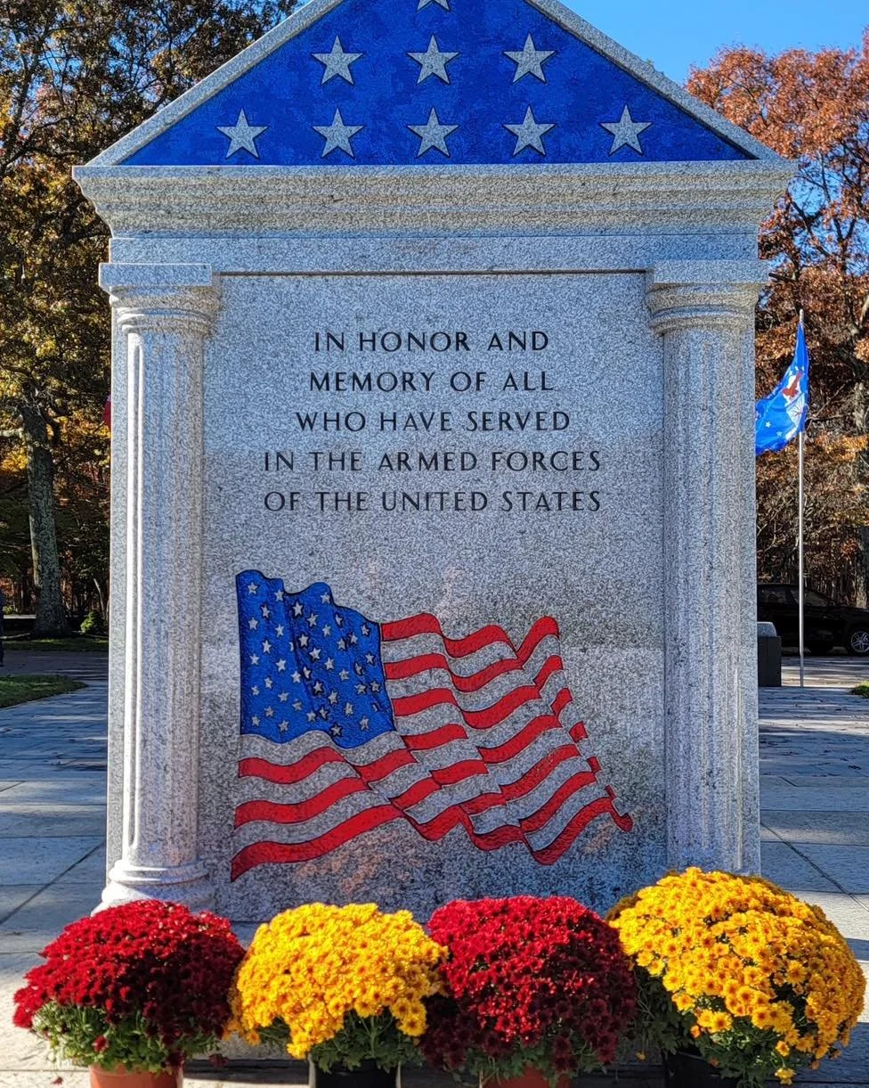 A memorial plaque with a blue triangular top decorated with white stars and a main gray stone panel inscribed with a tribute to those who served in the armed forces of the United States. The plaque features a painted American flag and is surrounded b