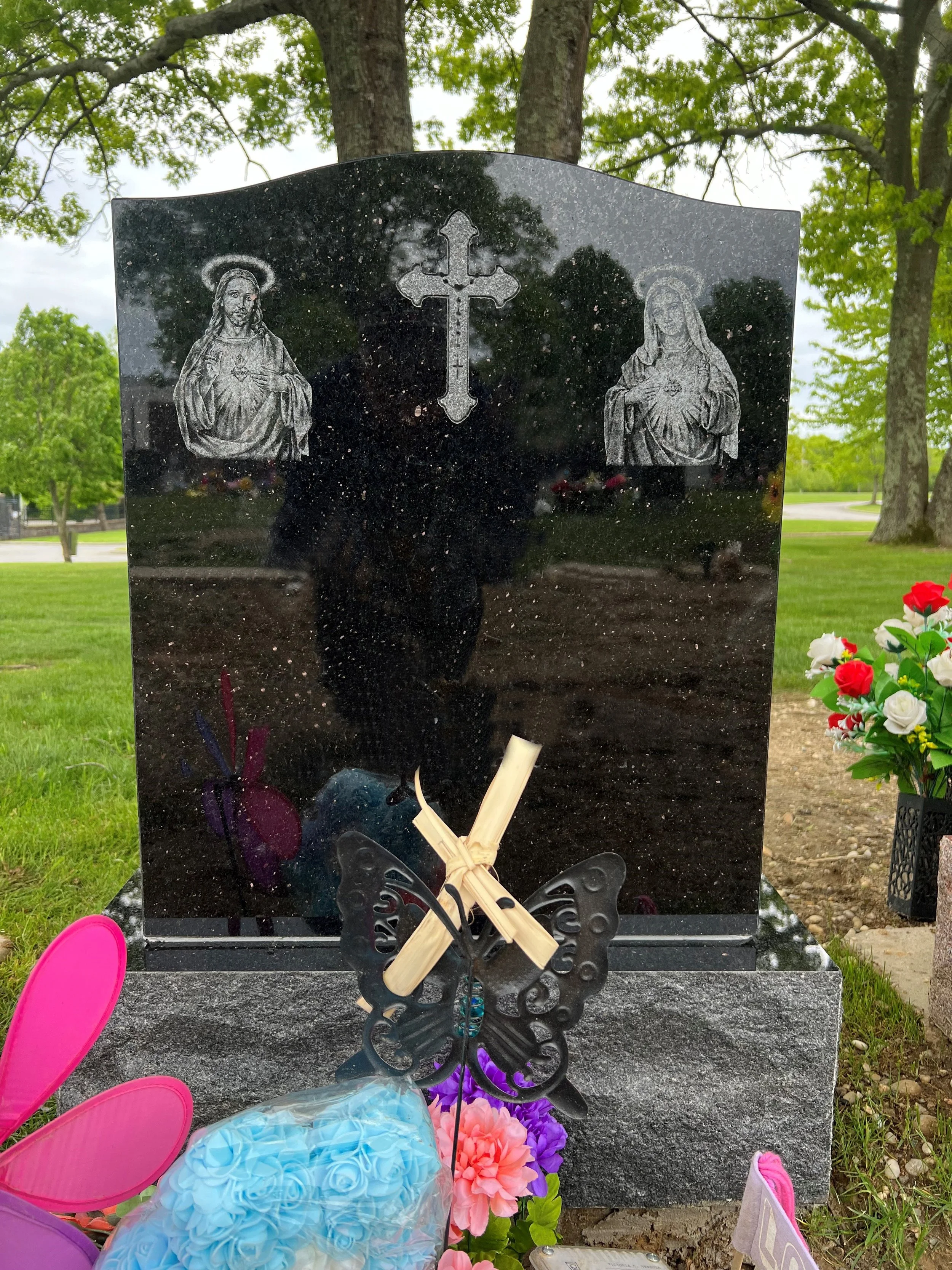 A black polished granite headstone with engraved images of Jesus Christ and the Virgin Mary, featuring halos and sacred hearts, in a cemetery with greenery and flowers nearby.