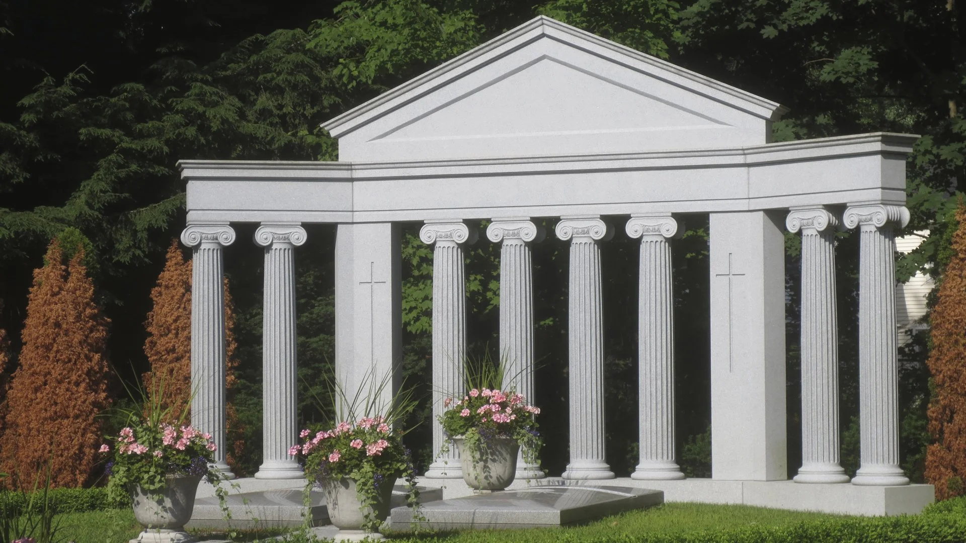 A white Greek-style temple display with columns and a triangular pediment, surrounded by pink and purple flowers, green shrubs, and trees.