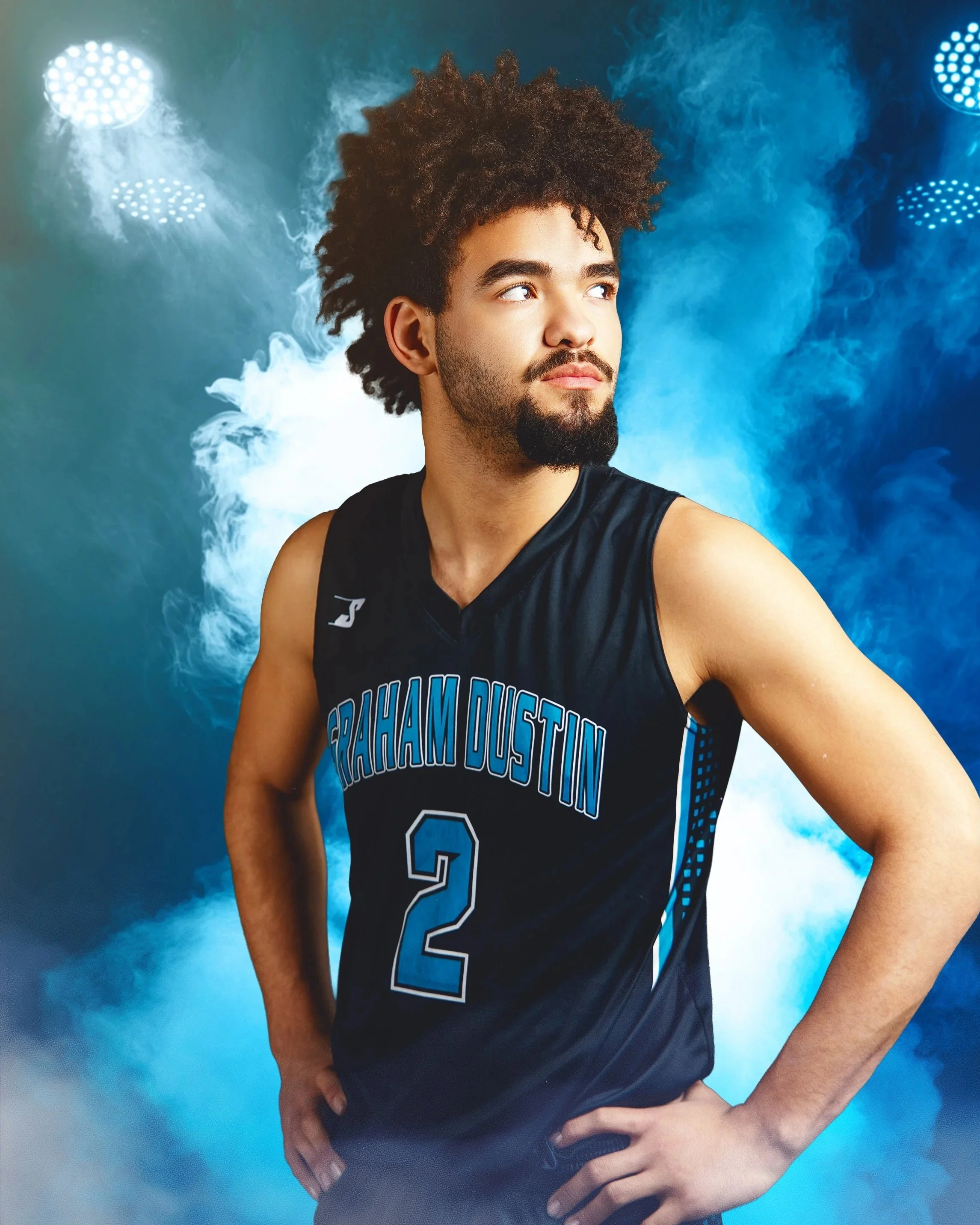 A male basketball player in a black jersey with the text 'GRAHAM DUSTIN' and the number 2, standing with his hands on his hips in front of a smoky blue background with bright lights.
