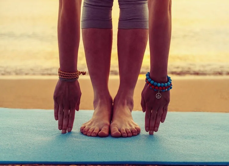 Person doing yoga stretch on blue mat at beach, wearing bracelets