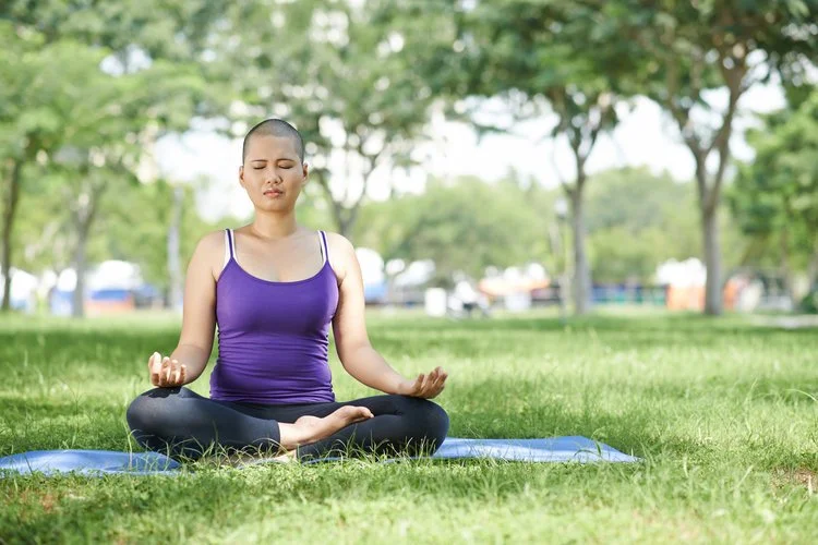 Person meditating outdoors on a yoga mat in a park, sitting cross-legged and wearing a purple tank top and black leggings.