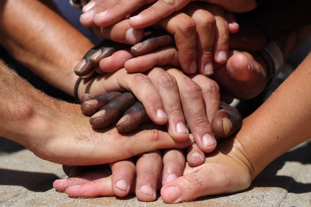 Close-up of multiple hands stacked together, displaying unity and teamwork on a sandy surface.