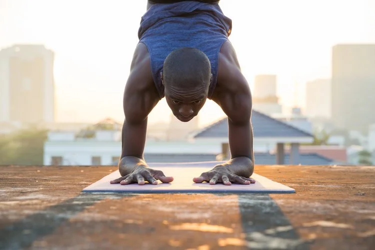 Person practicing a yoga pose on a rooftop with a city skyline in the background during sunrise or sunset.