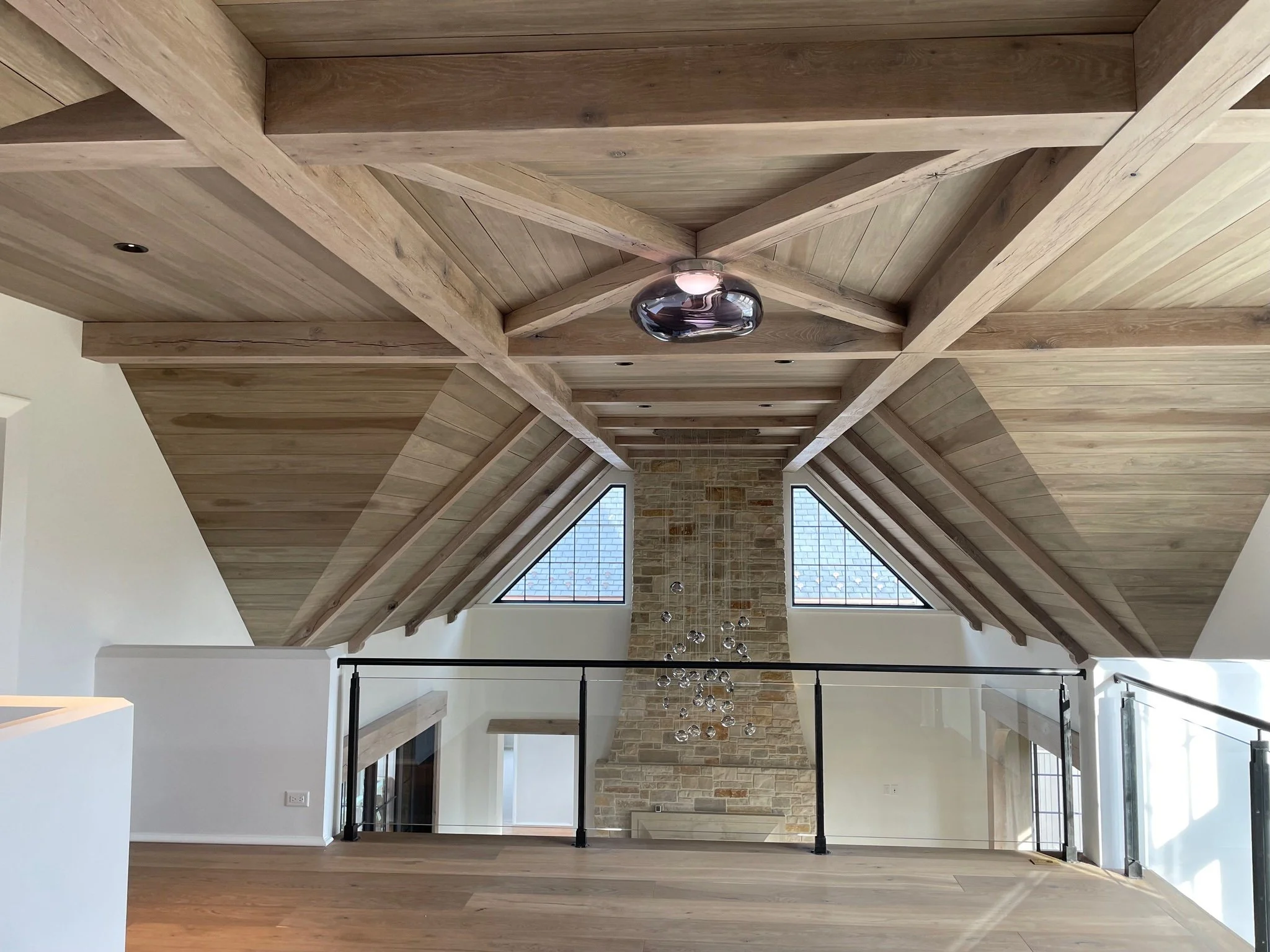 View of a high ceiling with wood beams and panels, a stone fireplace, triangular windows, and a modern chandelier hanging from the ceiling.