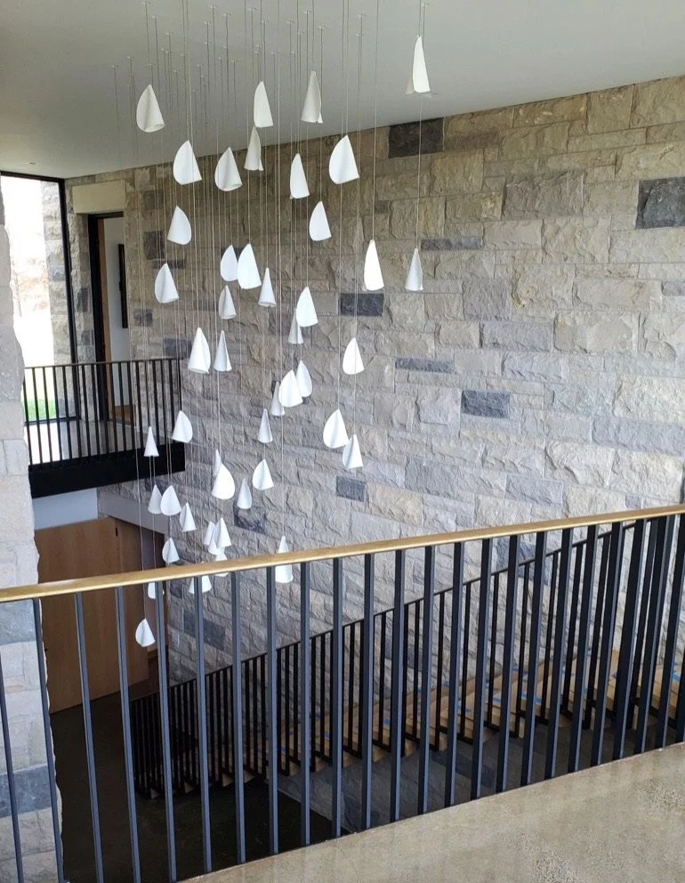 Interior view of a modern building with a stone wall, railing, stairs, and a hanging sculpture of white cone-shaped objects.