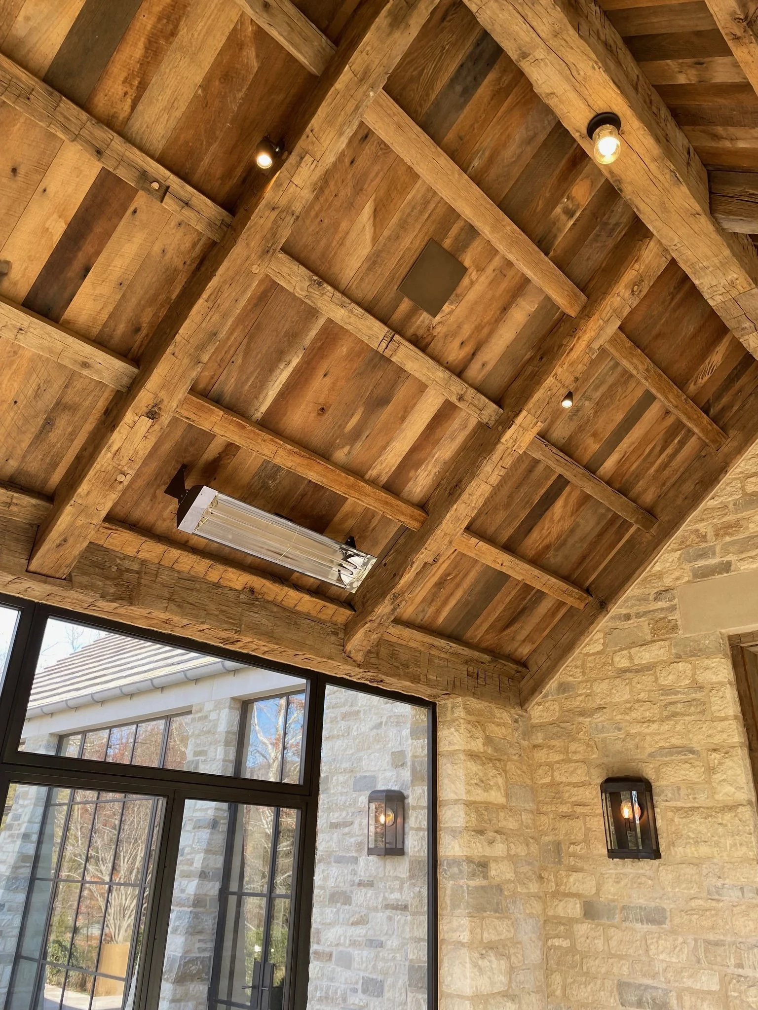 Wooden ceiling with exposed beams, ceiling lights, brick wall with lantern-style wall lights, large window with grid pattern showing outdoor trees.