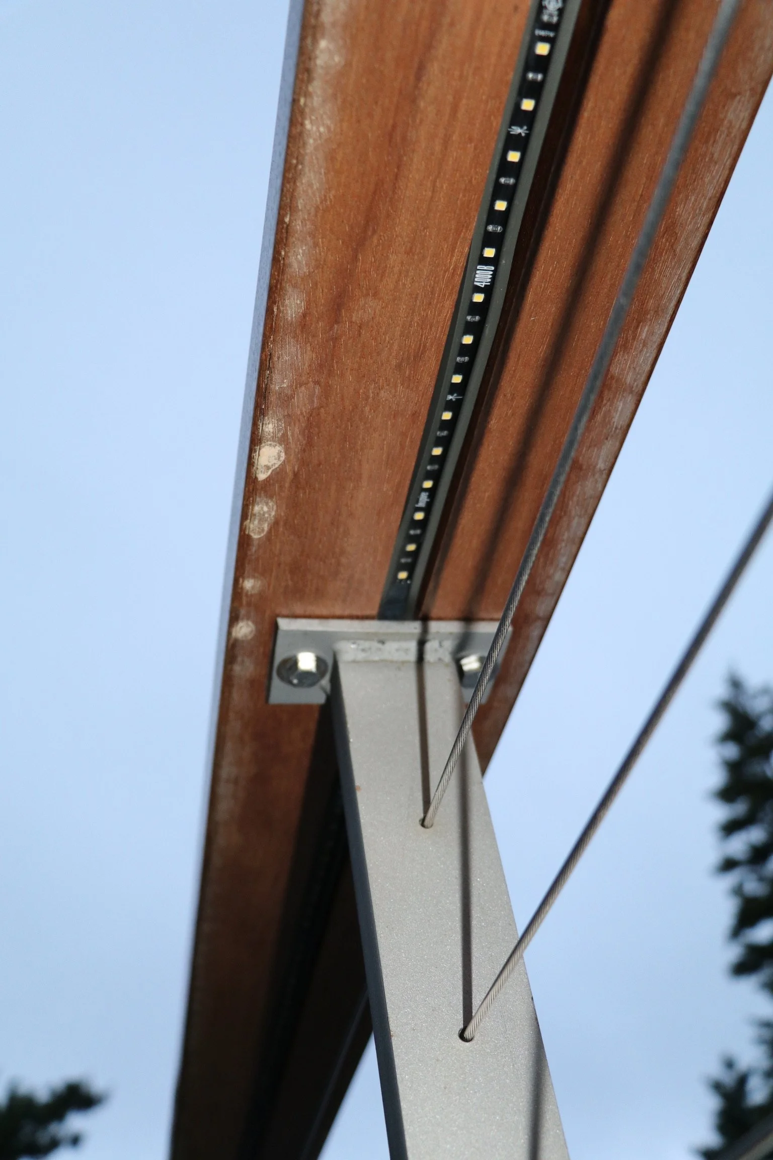 Close-up view of the underside of a wooden utility pole with LED lights and metal supports, against a clear blue sky with trees in the background.