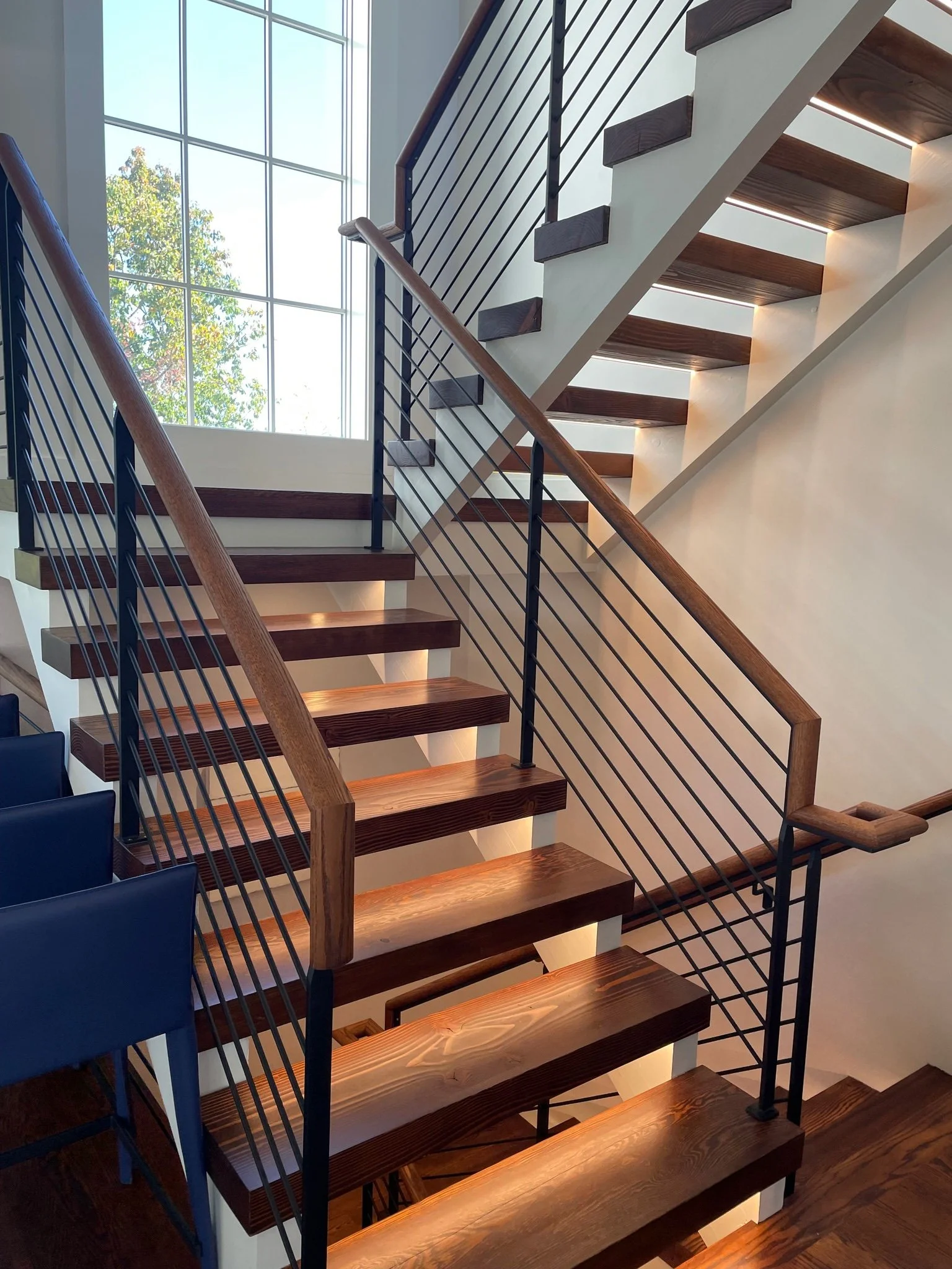 Interior view of a modern staircase with wooden steps, black metal railing, and a large window allowing natural light in.