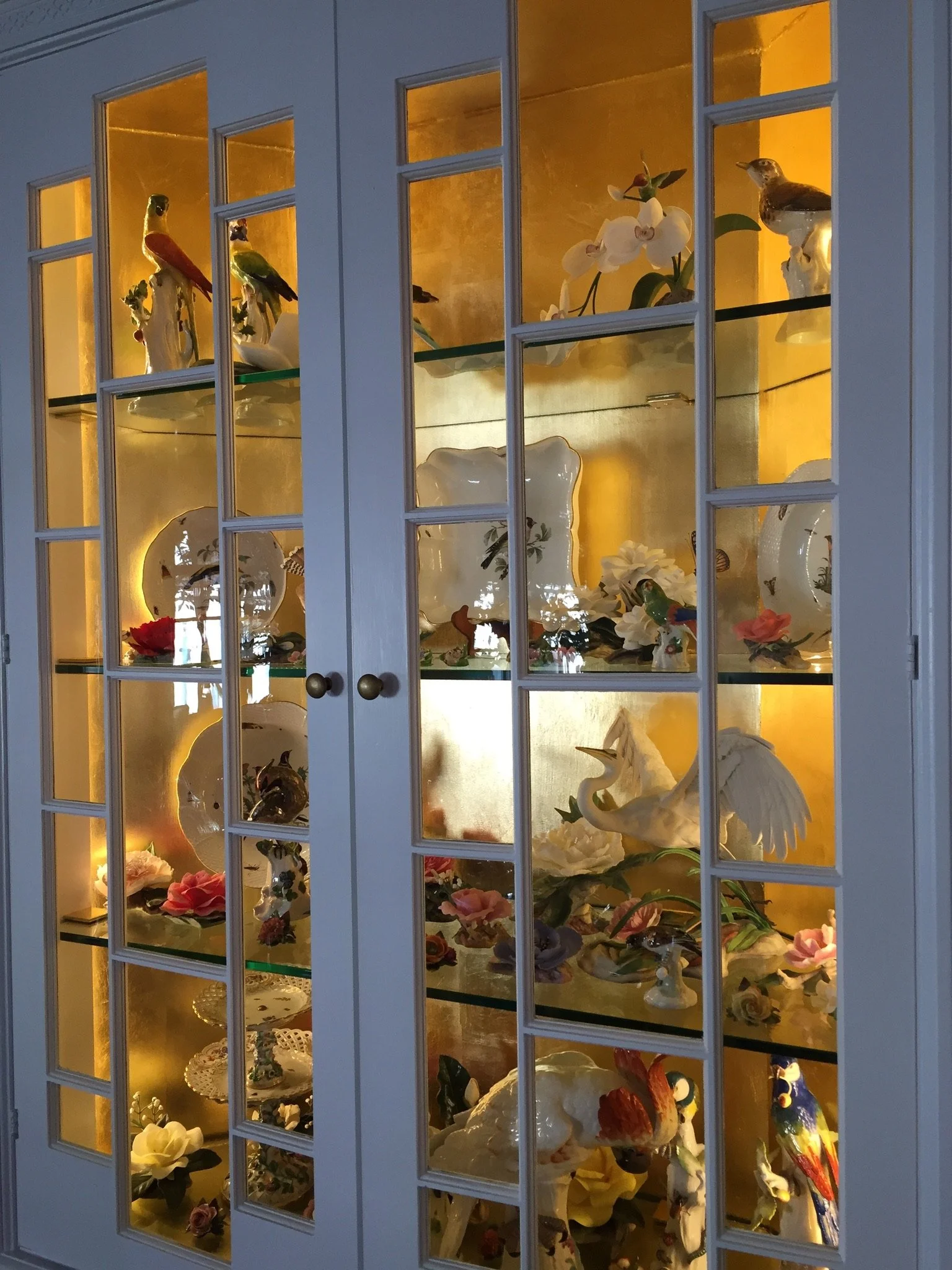 A glass display cabinet with a white frame and multiple shelves, illuminated from inside, showcasing various decorative porcelain figurines of birds, flowers, and other ornaments.