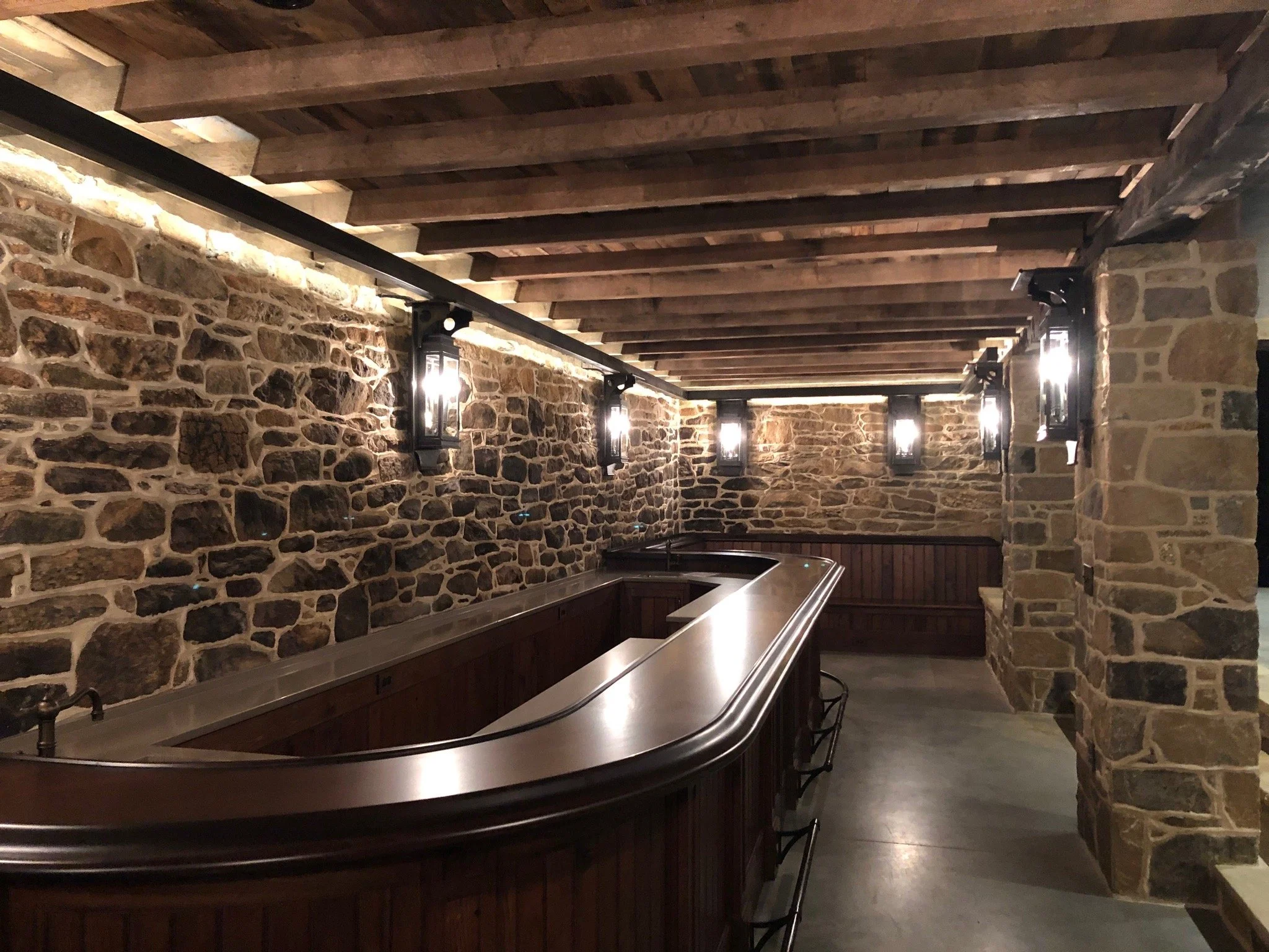 A rustic bar with a curved wooden counter, stone walls, and black lantern-style wall sconces lighting the space with a warm, inviting glow.