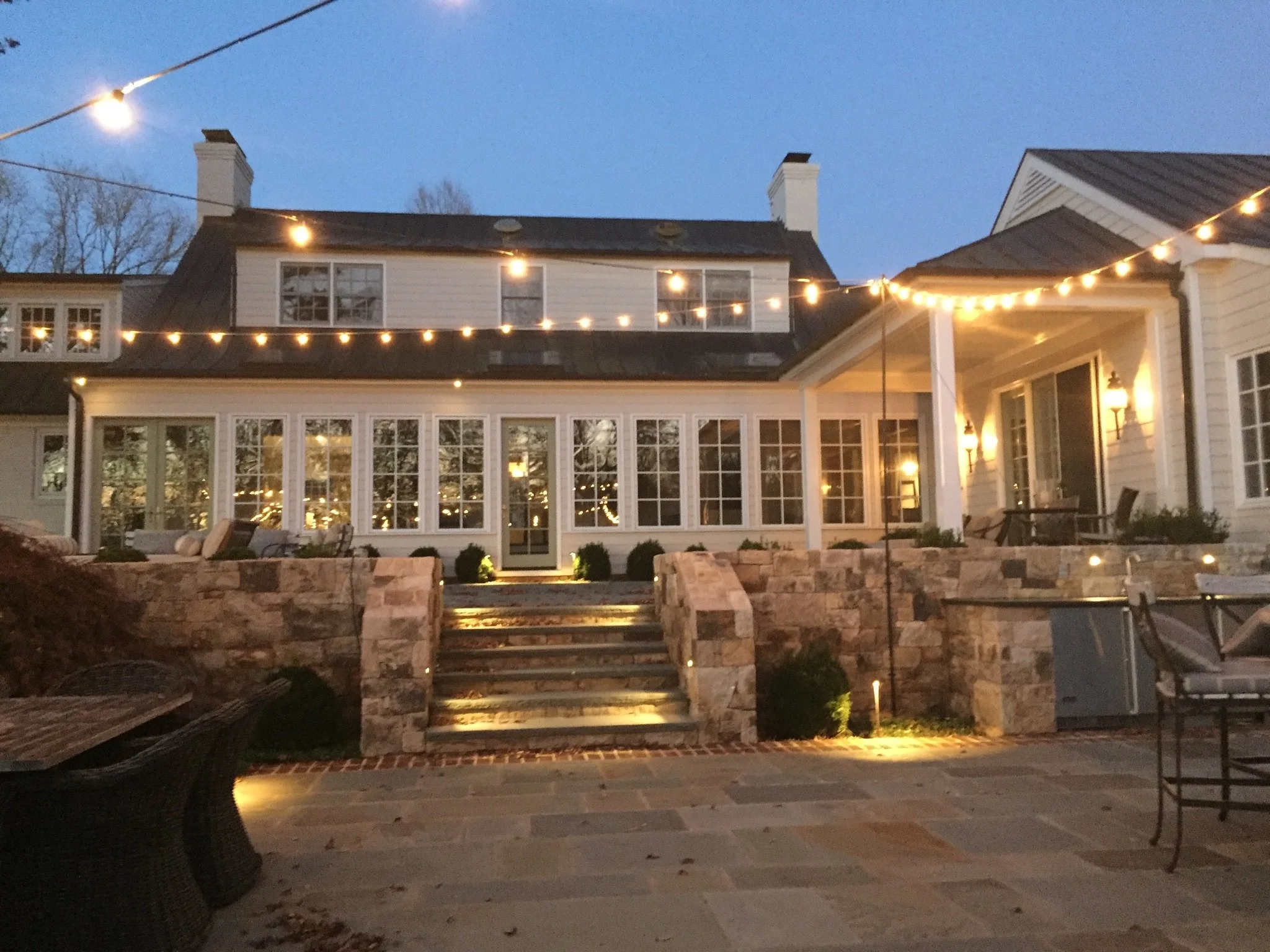 Backyard patio with stairs, string lights, outdoor seating, and house with large windows illuminated at dusk.