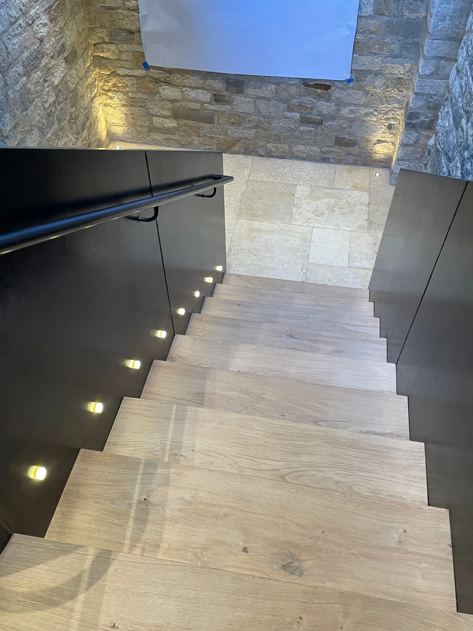 Top-down view of a staircase with light-colored wooden steps, black side panels, and small built-in lights along the side. At the top, there's a stone wall with warm lighting, and a piece of paper or sign taped to the wall.