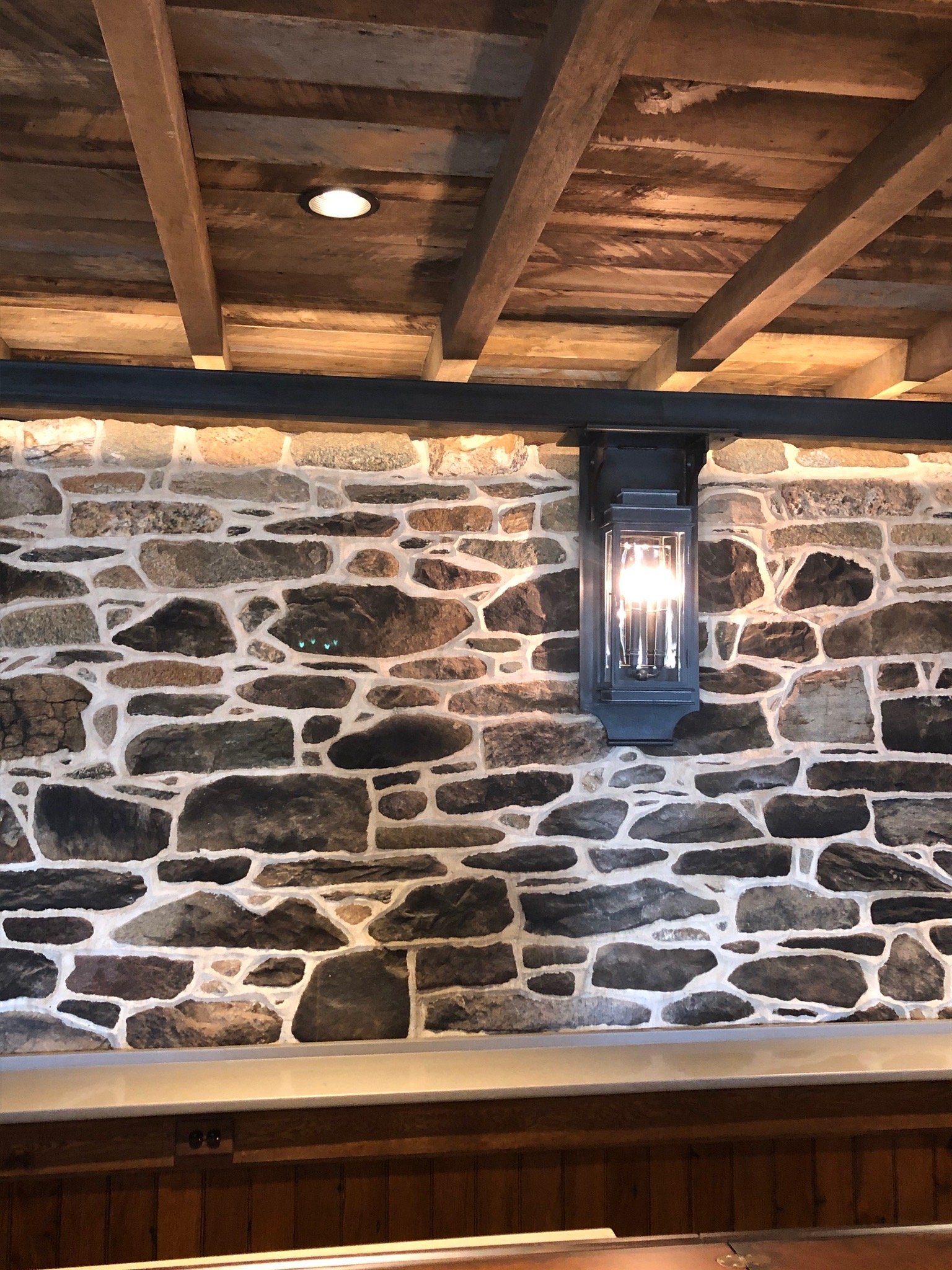 Interior wall with stone masonry, wooden ceiling beams, and an outdoor-style wall lantern.